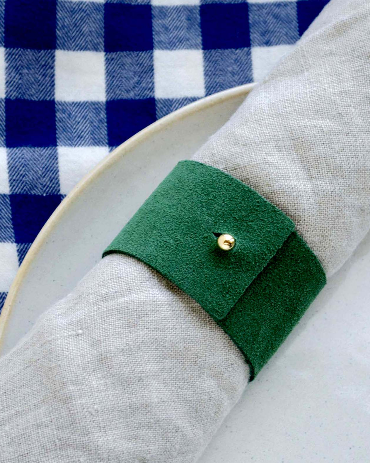 Moss green suede napkin ring with gold stud on a stoneware plate and a blue gingham table cloth for Christmas.