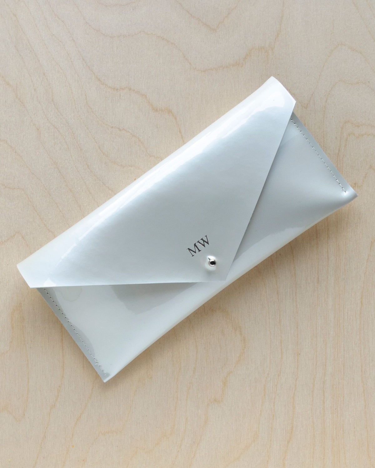 A white pearlescent leather clutch purse with a minimalist envelope design, displayed against a wooden background.