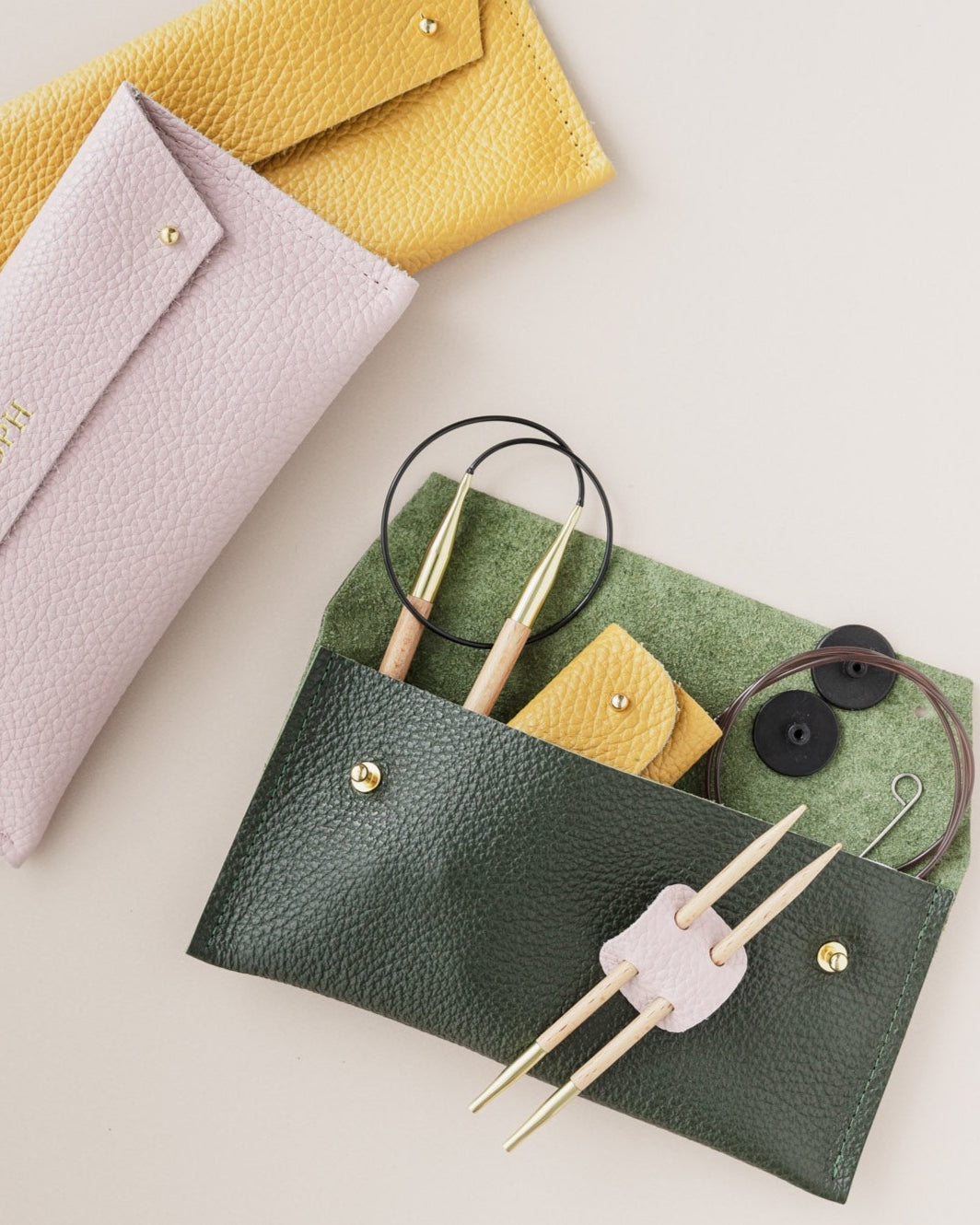 Yellow, pink and green sewing cases sit on a cream background. The green pouch shows circular knitting needles and notions inside it.