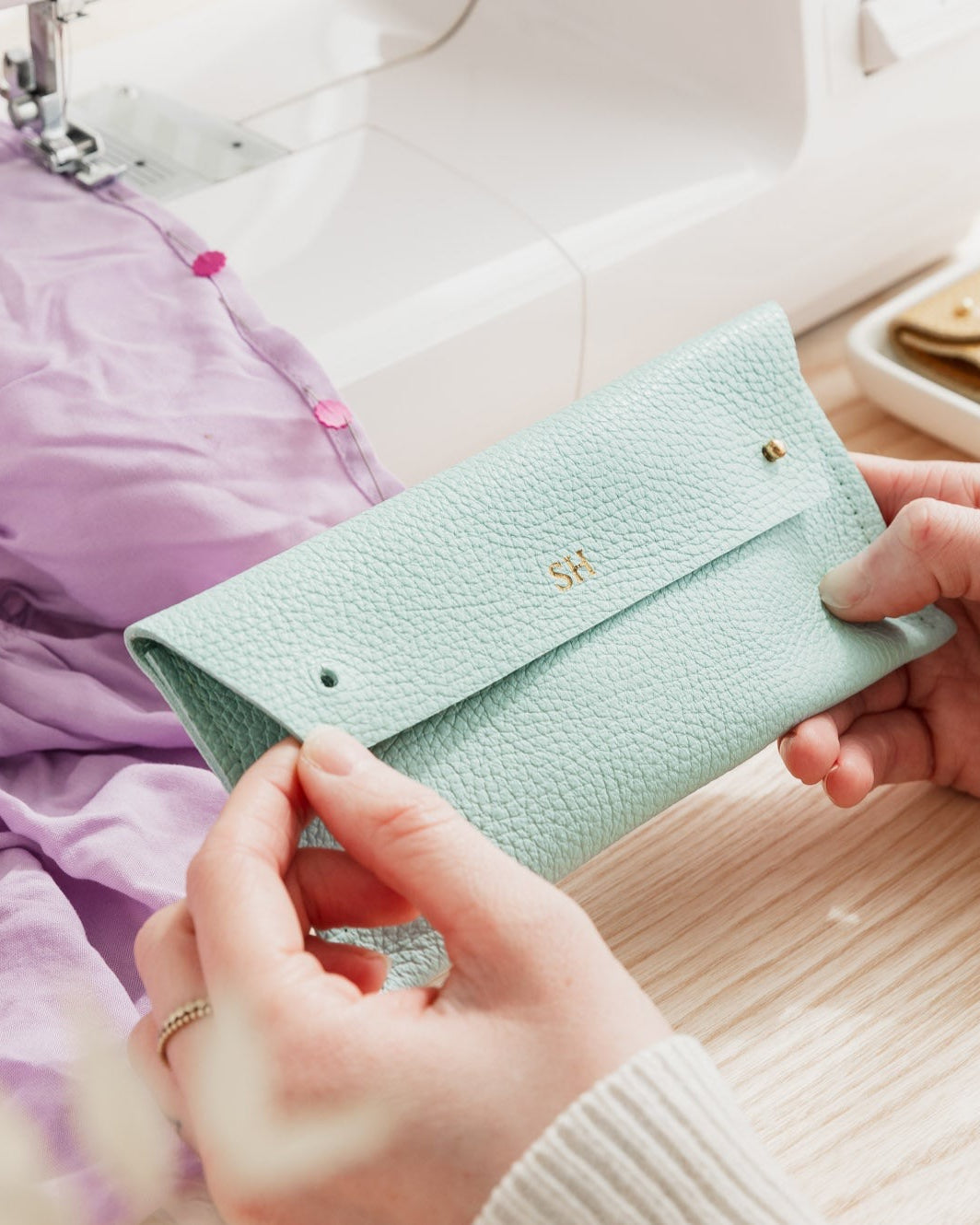 Sewer holds light turquoise leather sewing case with two metal studs while she sews lilac dress. The leather pouch is personalised with the initials SH.
