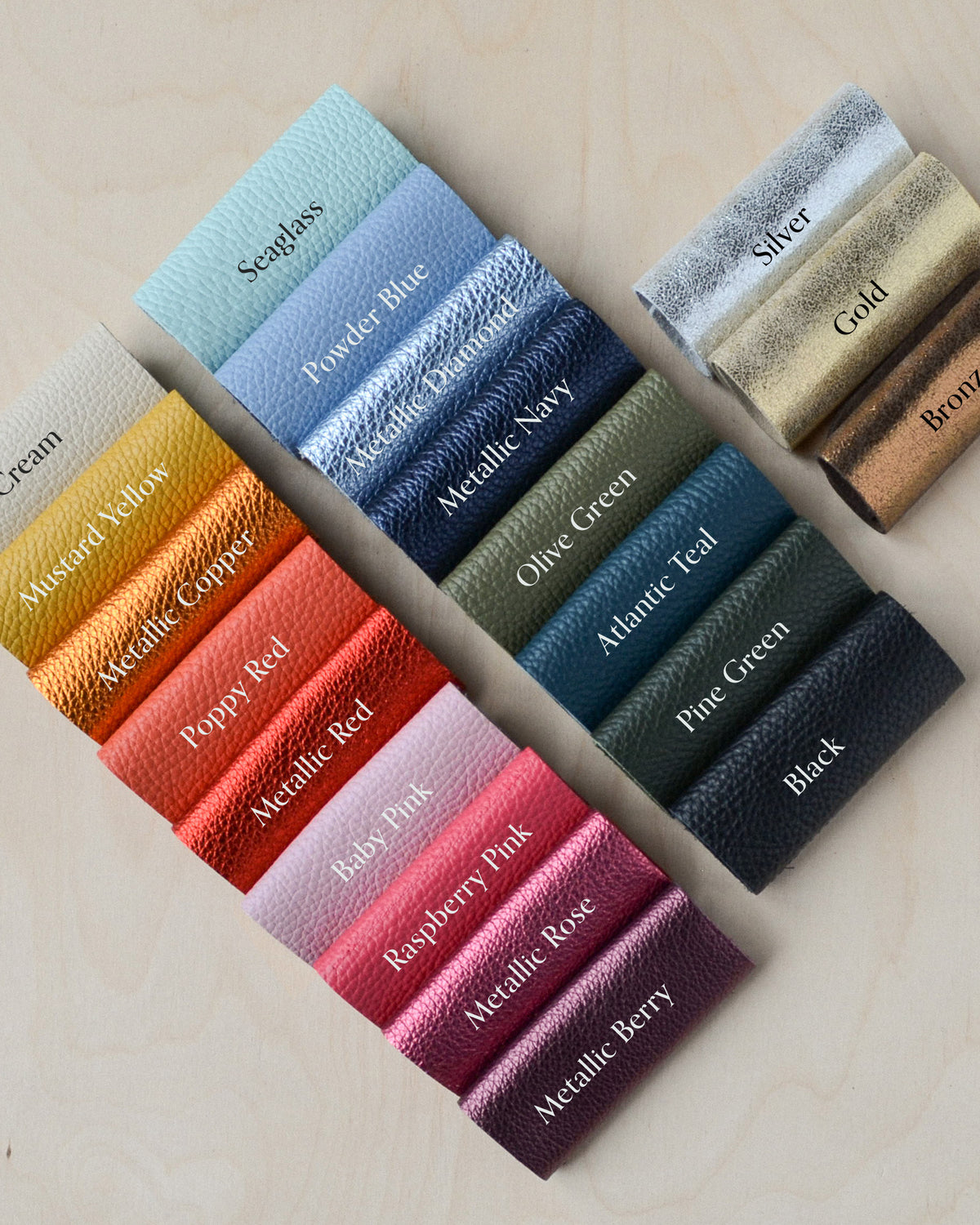 A set of Italian leather color swatches arranged on a light wood background, showing various colors and metallic finishes including Cream, Mustard Yellow, Powder Blue, Metallic Navy, Silver, Gold, Bronze, Olive Green, Atlantic Teal, Pine Green, Black, Metallic Rose, and Metallic Berry.