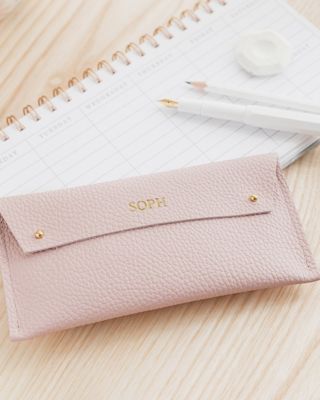 Pink leather pencil case sits on wooden desk with white minimalist pens. The pink leather case is personalised with the name Soph.