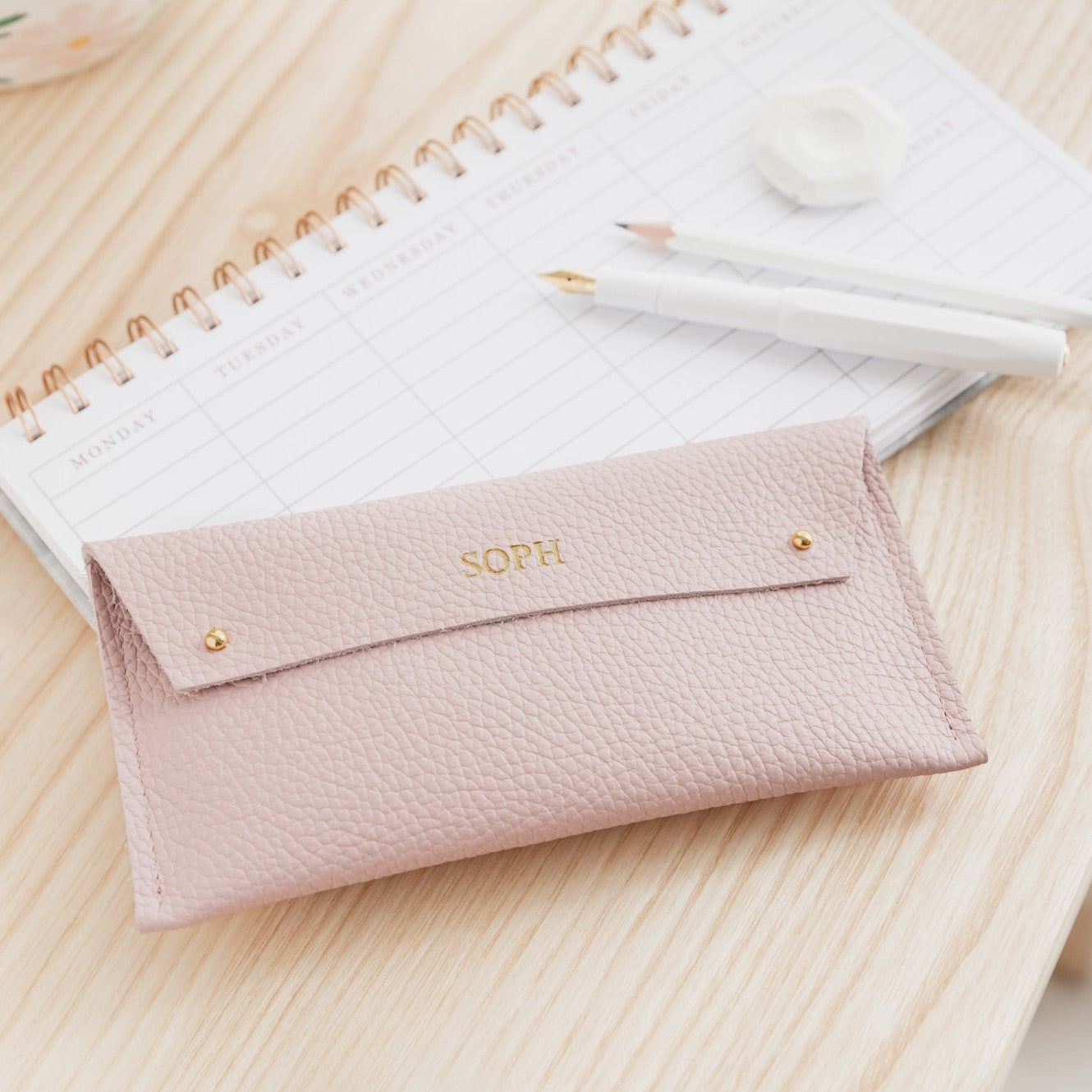Pink leather pencil case sits on wooden desk with white minimalist pens. The pink leather case is personalised with the name Soph.