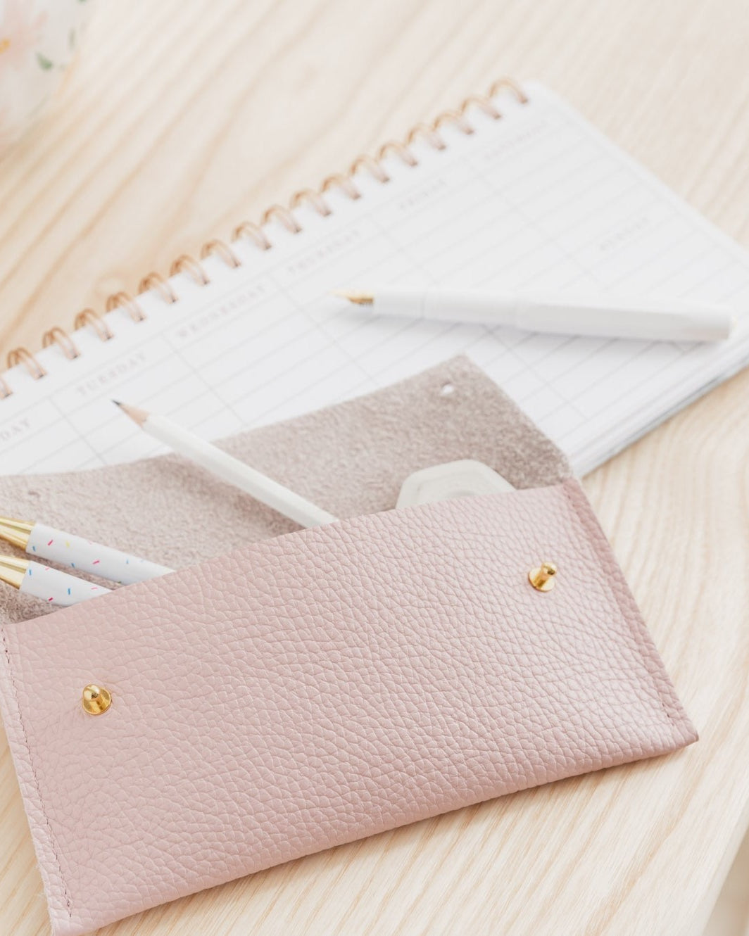 Open pink leather pen case with white stationery and pens displayed in the case. Sits on top of a white calendar on a pine wooden desk.