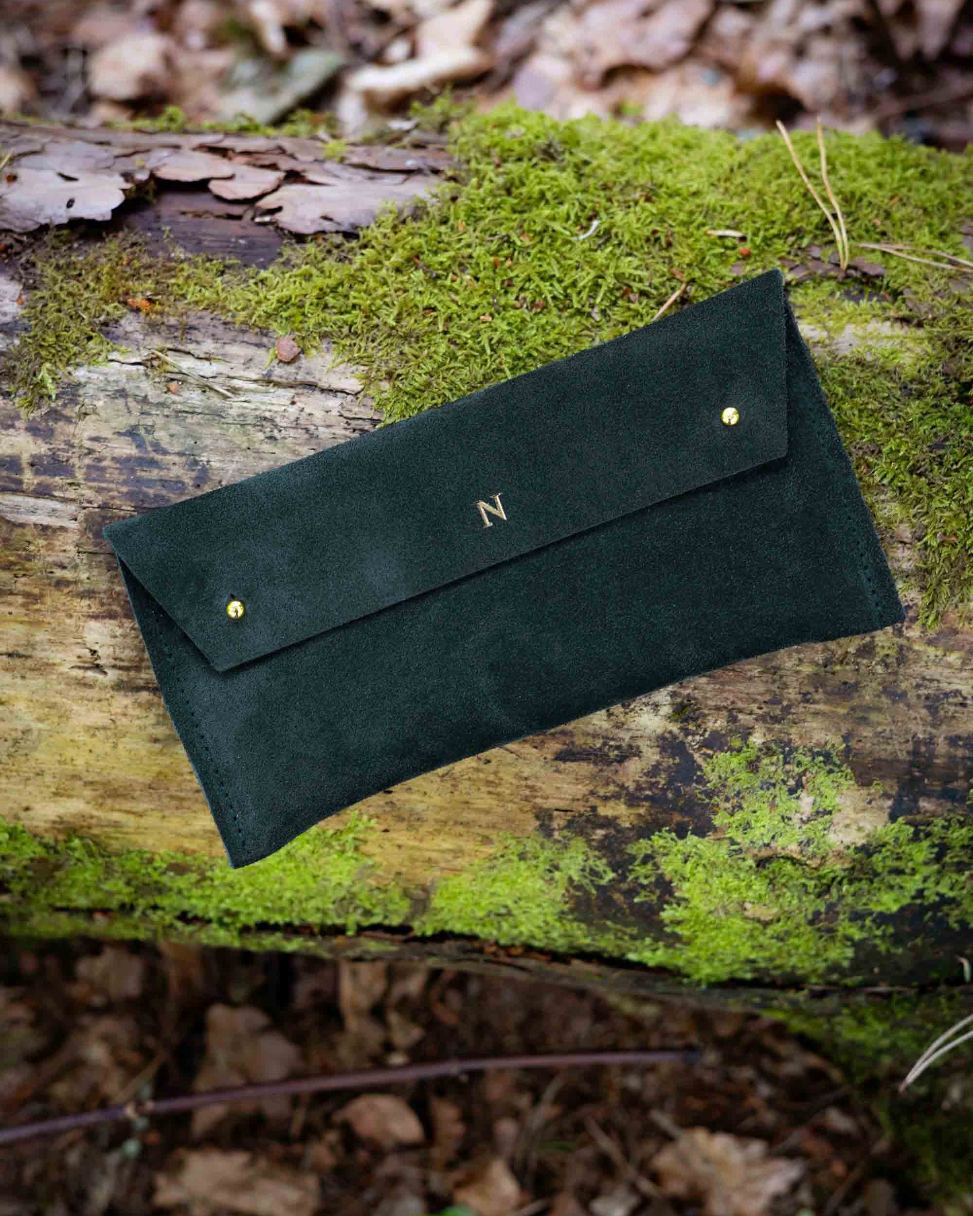 A dark forest green suede clutch or wallet with gold-tone studs and a small monogram 'N' on the flap, resting on a mossy log in a forest setting.