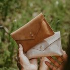 Brown suede and gold leather envelope purse held in ladies hands in a field in summer. The purse is personalised with the letter B in gold.