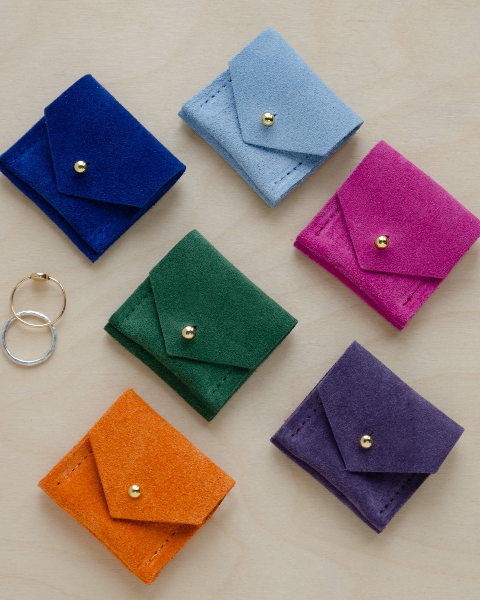 Six of the Wedding Ring Pouches in suede. The colours are, Ultramarine Blue, Pale Blue, Moss Green, Fuchsia Pink, Clementine Orange and Grape Purple.