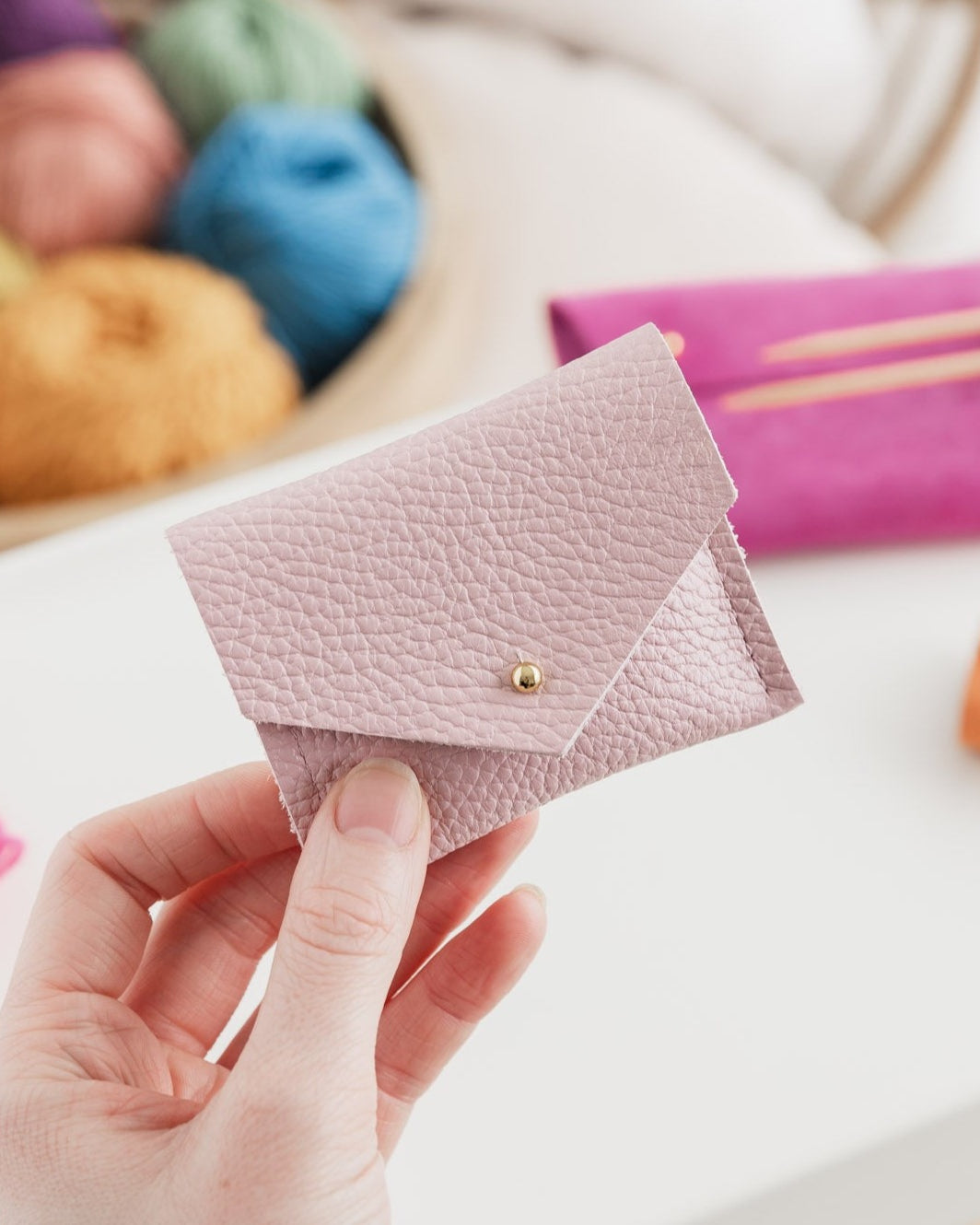 Baby pink leather pouch held in hands in front of balls of colourful wool and knitting accessories.