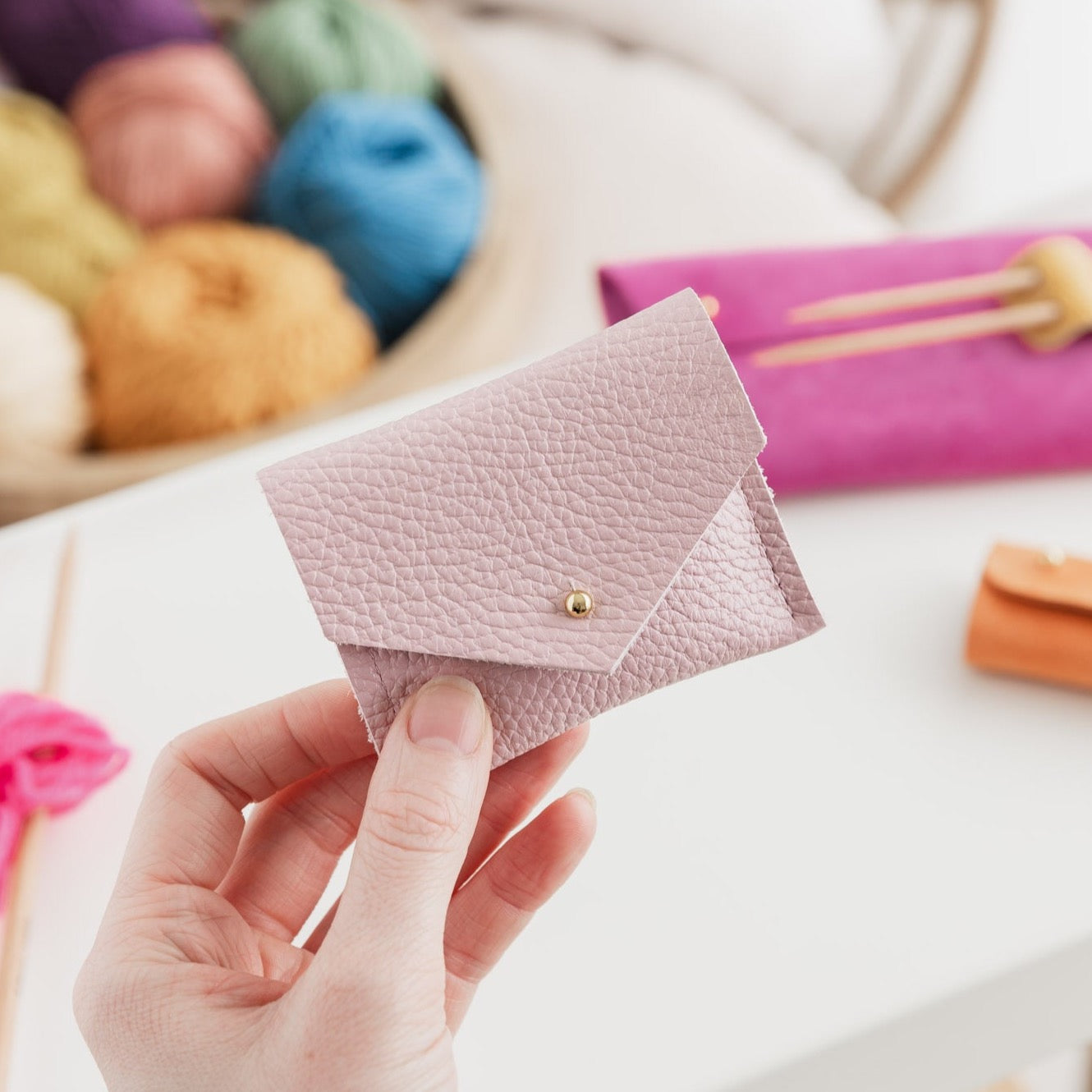 Baby pink leather pouch held in hands in front of balls of colourful wool and knitting accessories.