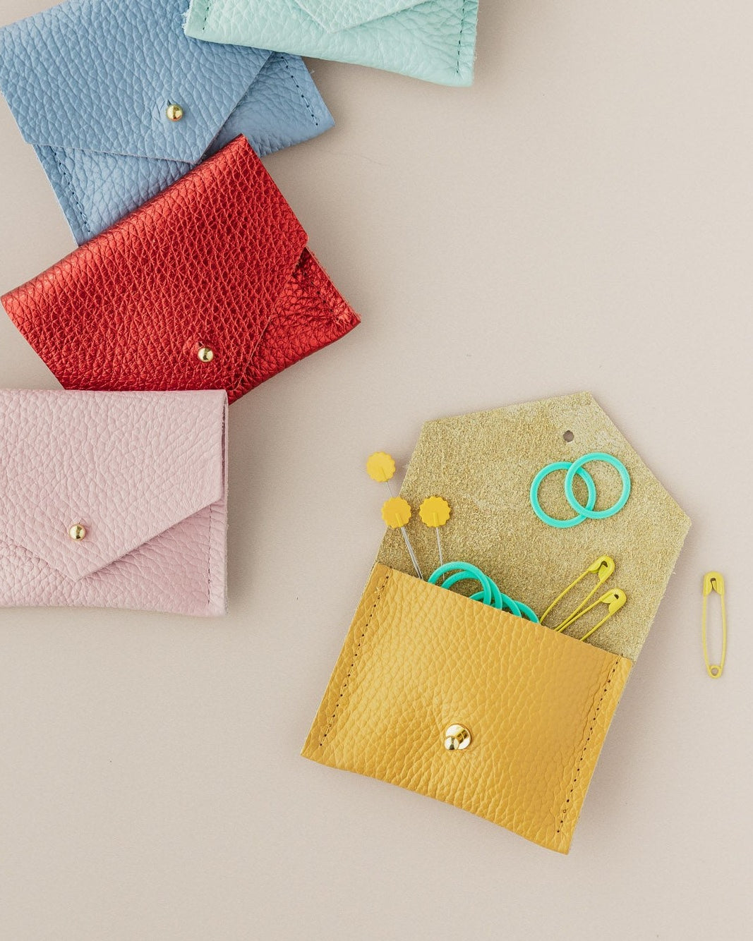 Leather pouch in yellow, baby pink, metallic red, powder blue and pale turquoise. The yellow pouch is open with sewing and knitting notions coming out of the pouch.