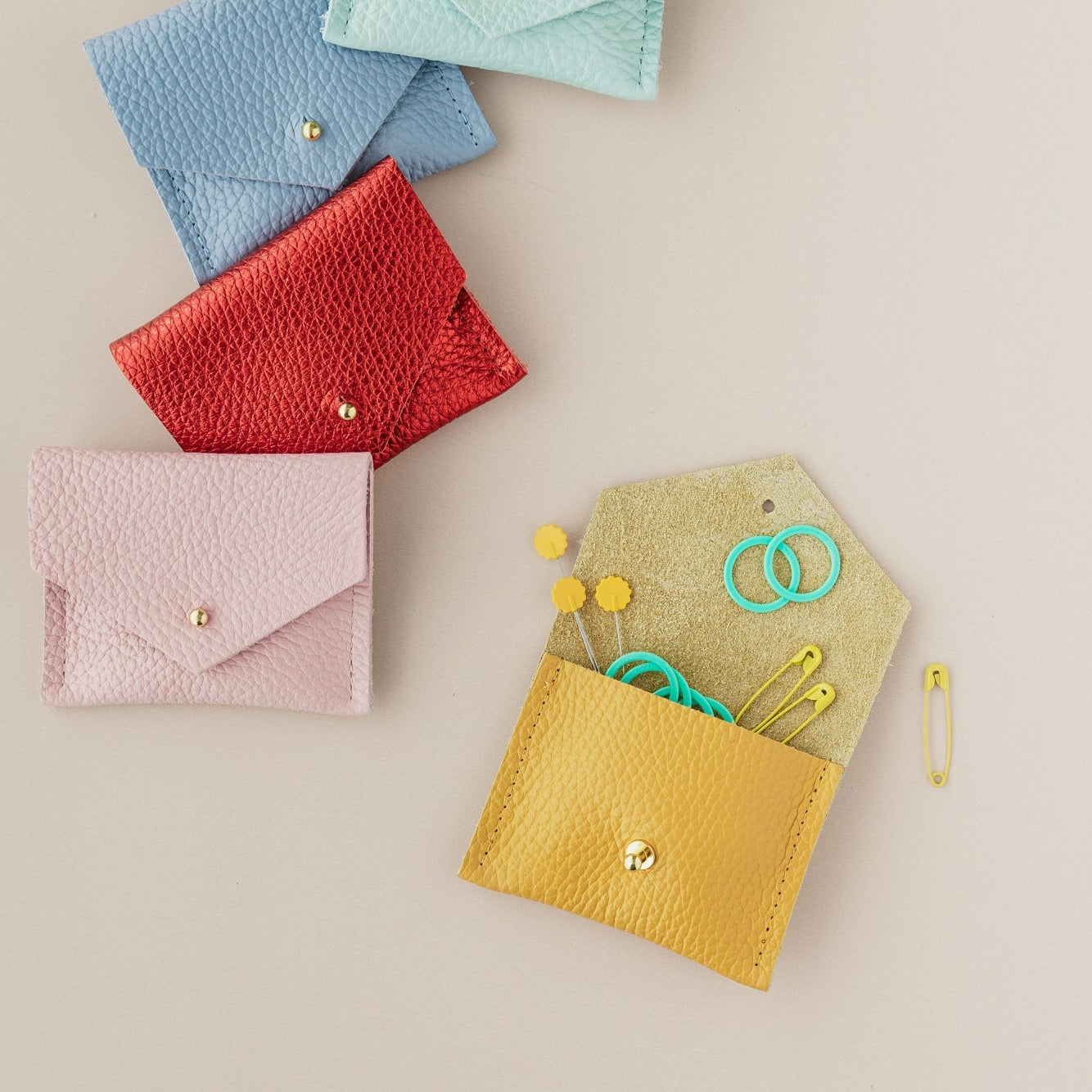 Leather pouch in yellow, baby pink, metallic red, powder blue and pale turquoise. The yellow pouch is open with sewing and knitting notions coming out of the pouch.