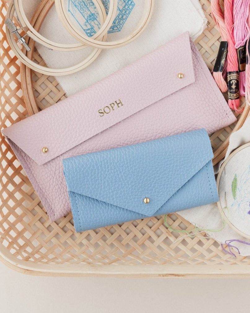Pink and blue sewing cases sit amongst embroidery threads and hoops in a wicker basket. The pink pouch is personalised with the name Soph in gold.
