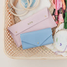 Pink and blue sewing cases sit amongst embroidery threads and hoops in a wicker basket. The pink pouch is personalised with the name Soph in gold.