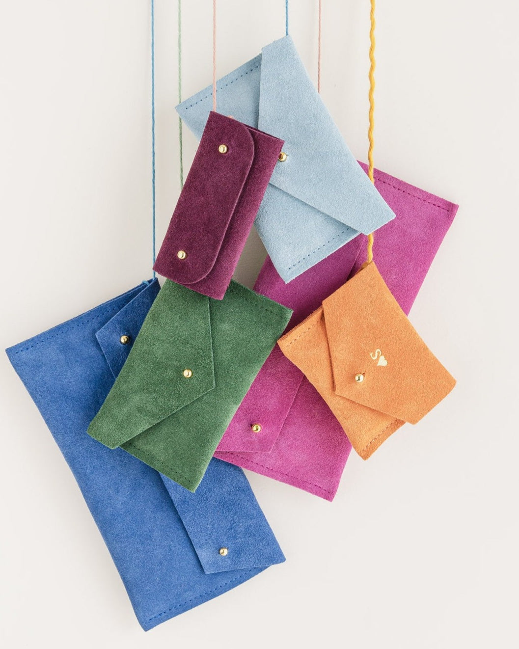 A group of pouches hanging from thread, the colours include: Plum, Pale Blue, Moss Green, Orange, Ultramarine and Fuchsia pink.