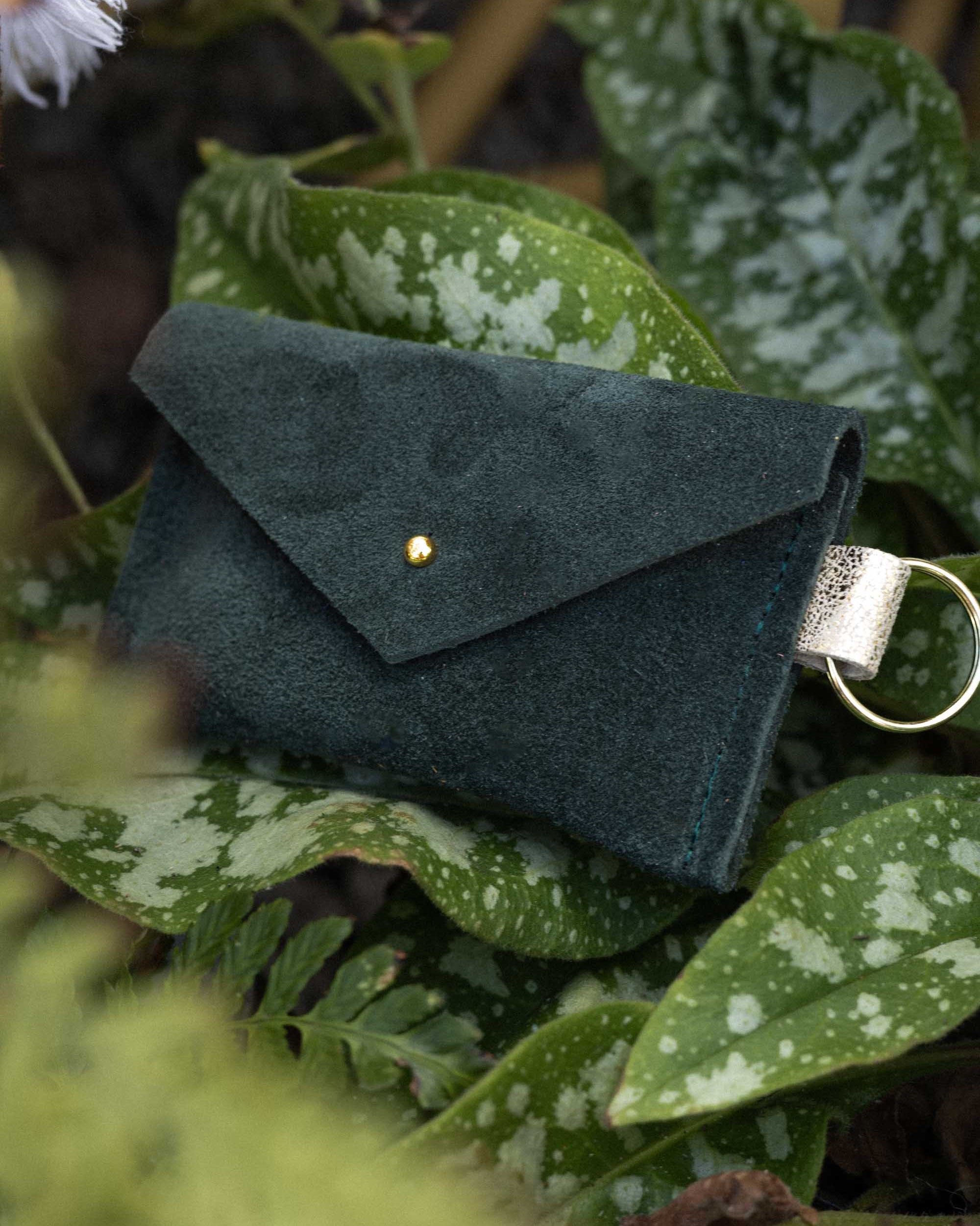 A small deep forest green coloured suede envelope-style keyring pouch with a gold button, nestled amongst some leaves in nature.