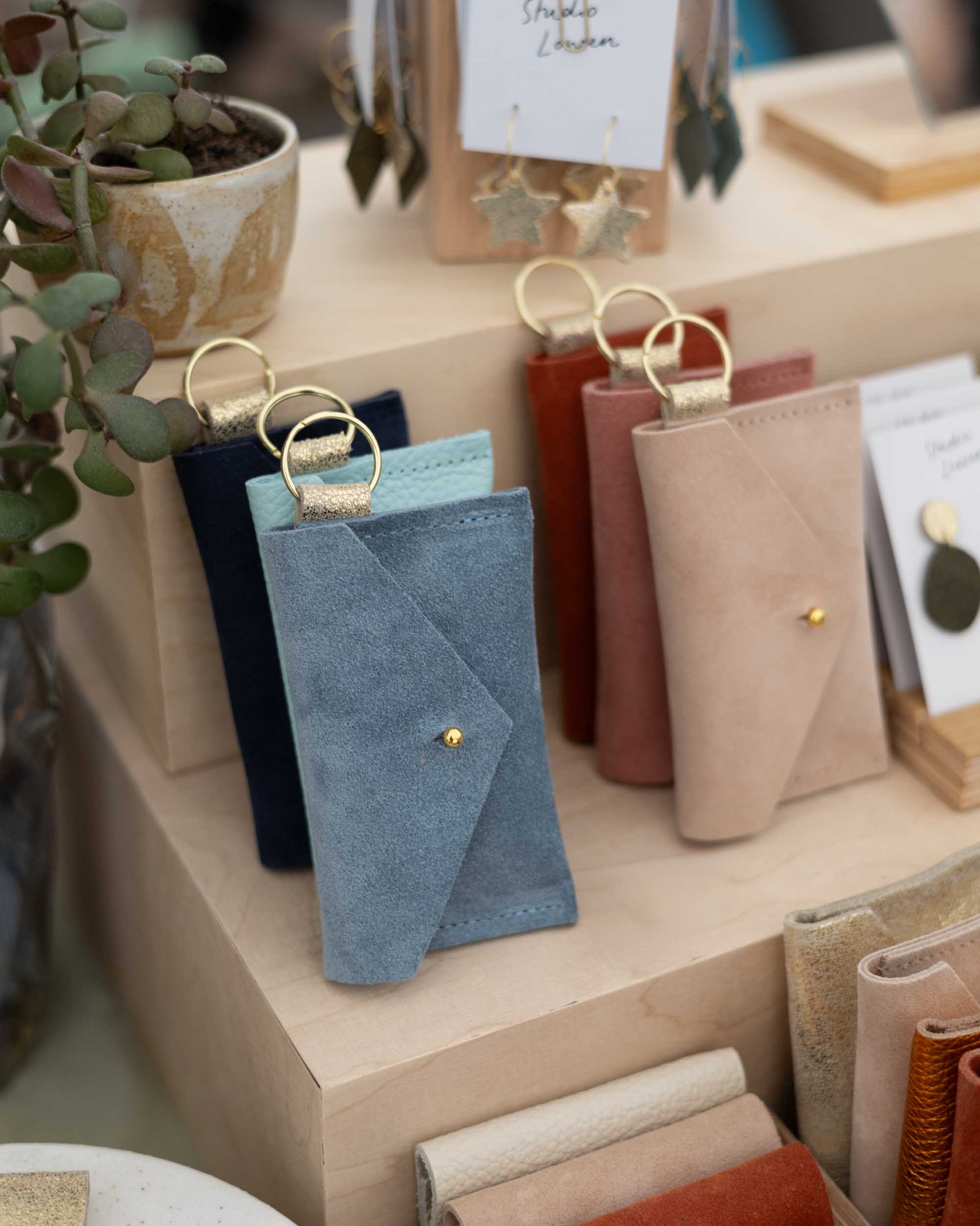 A photo of a retail display featuring suede pouches in different styles and assorted colours on a light wood grain shop display, with a small plant and a hanging display of leather earrings.
