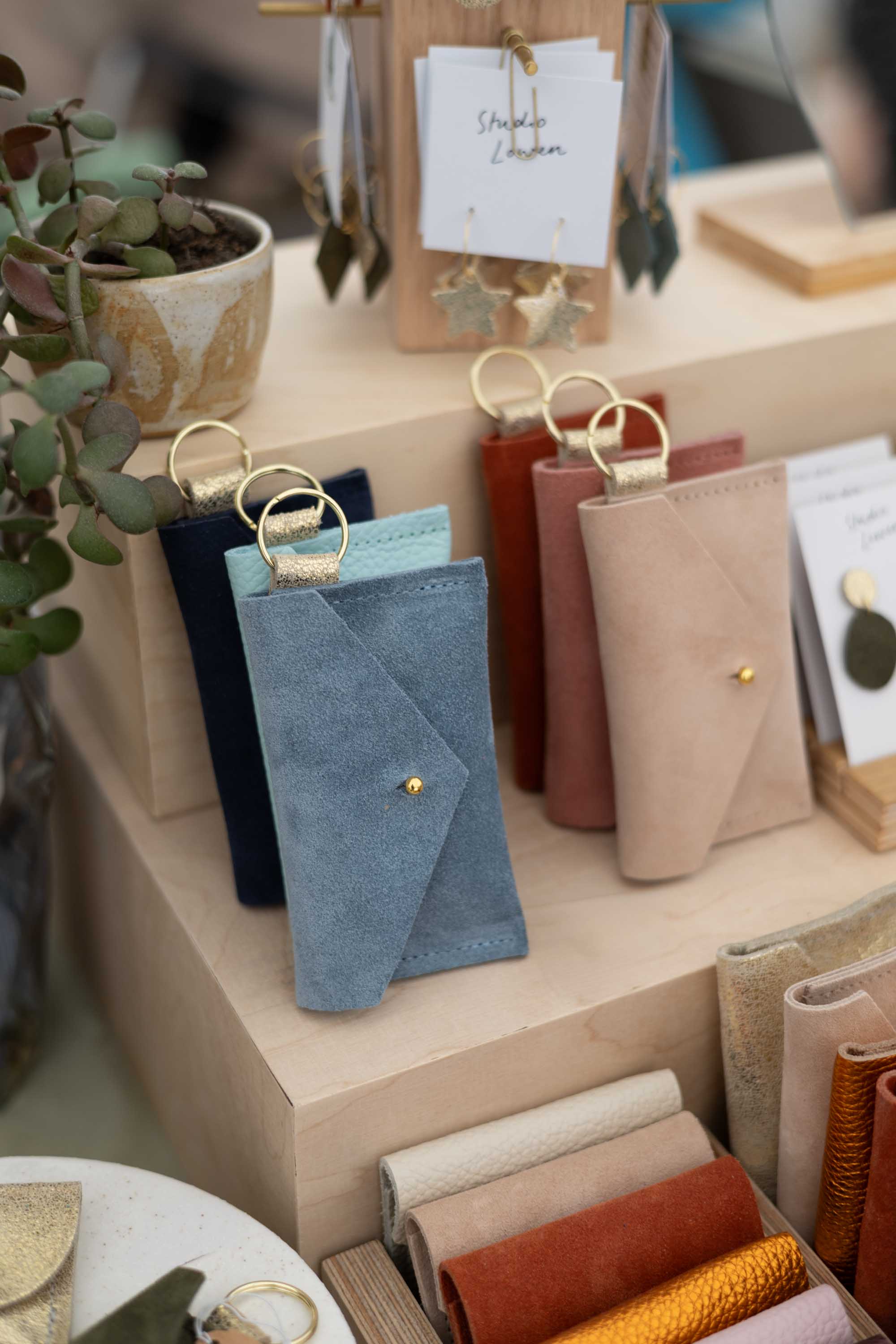 A photo of a retail display featuring suede pouches in different styles and assorted colours on a light wood grain shop display, with a small plant and a hanging display of leather earrings.