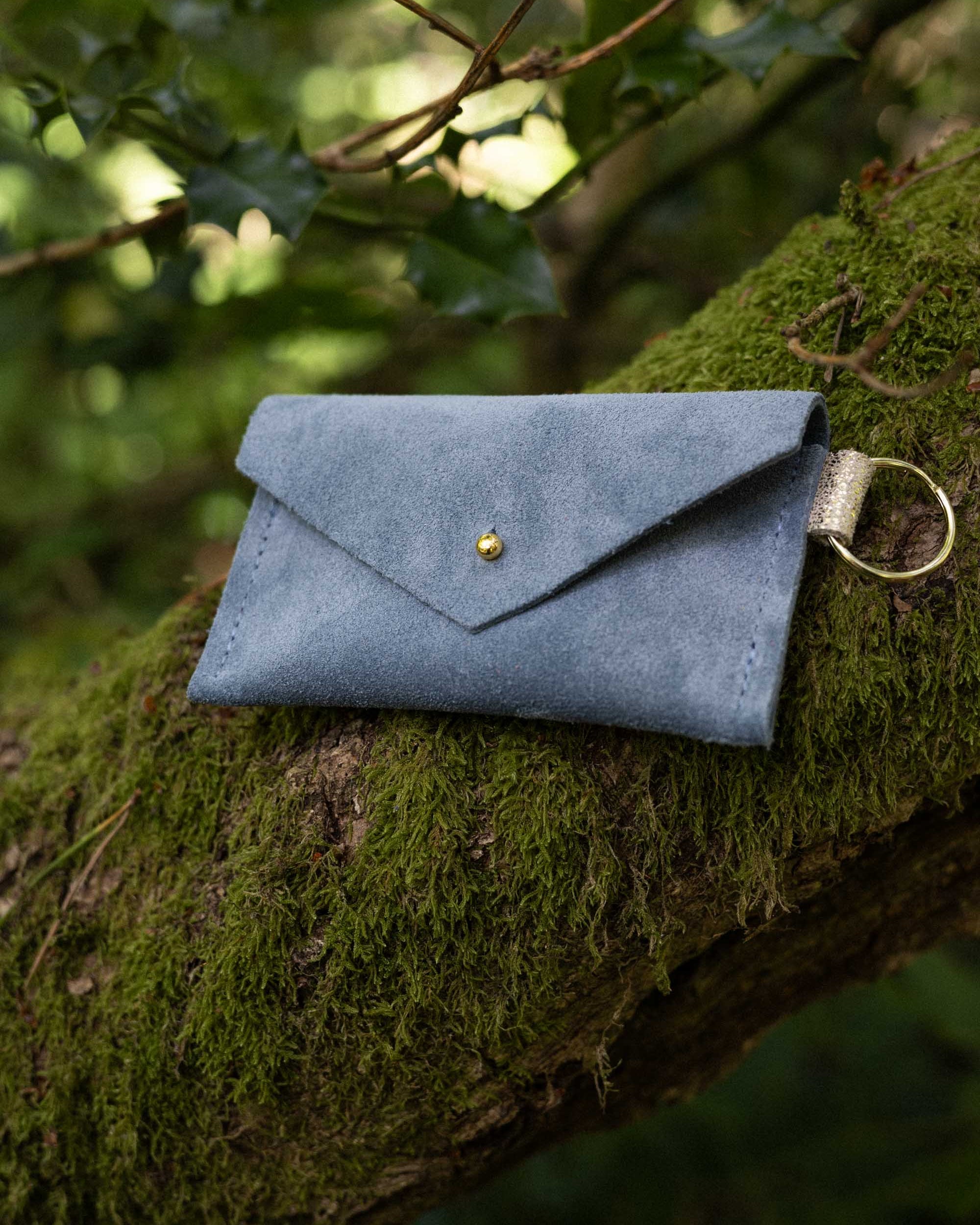 A small dusky blue coloured suede envelope-style keyring pouch with a gold button, resting on a tree branch in a forest.