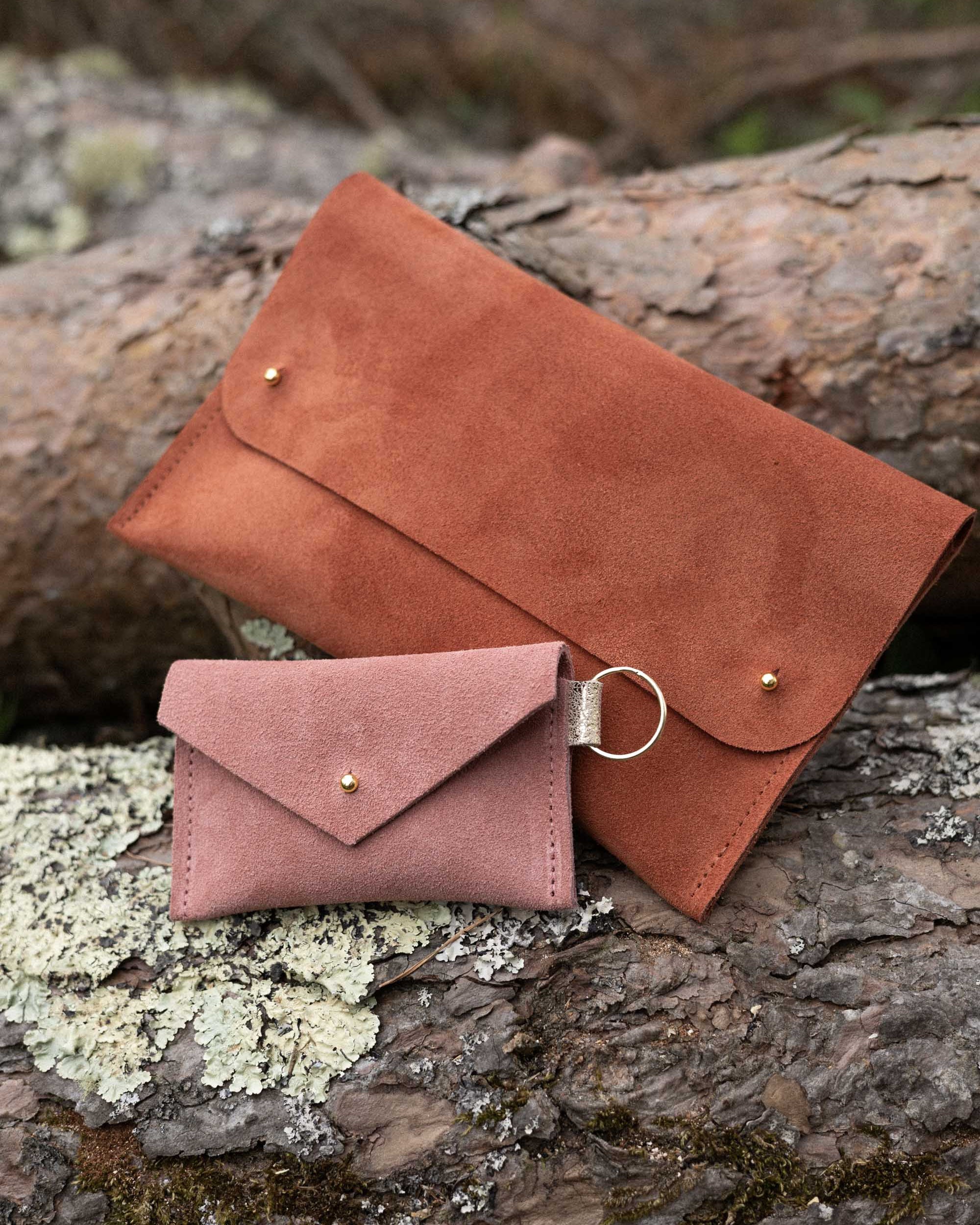 A small dusky rose coloured suede envelope-style keyring pouch with a gold button, resting on a light wood grain surface, alongside a rust coloured suede clutch bag.