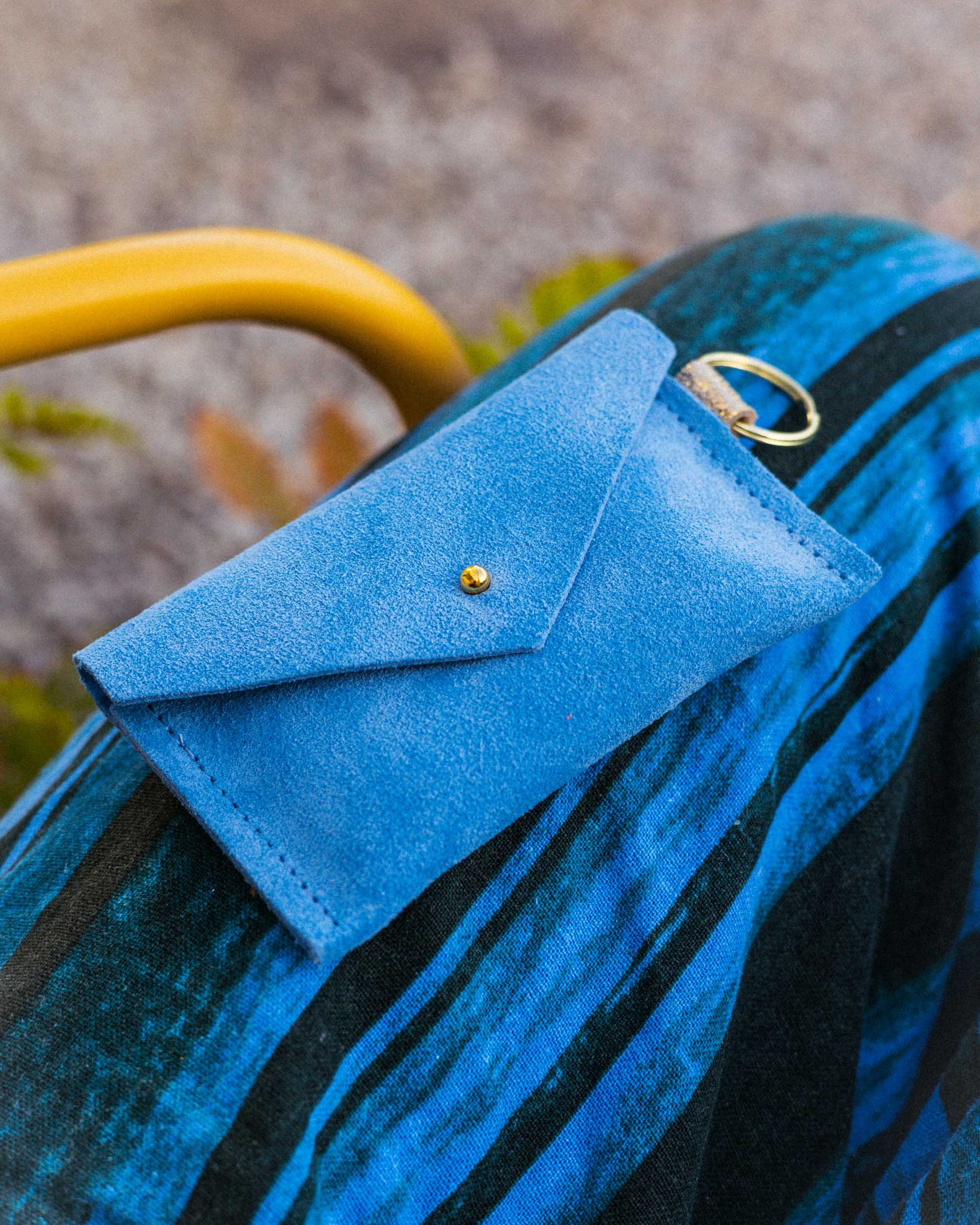 A small sky blue coloured suede envelope-style keyring pouch with a gold button, resting on a blue and black striped patterned fabric.