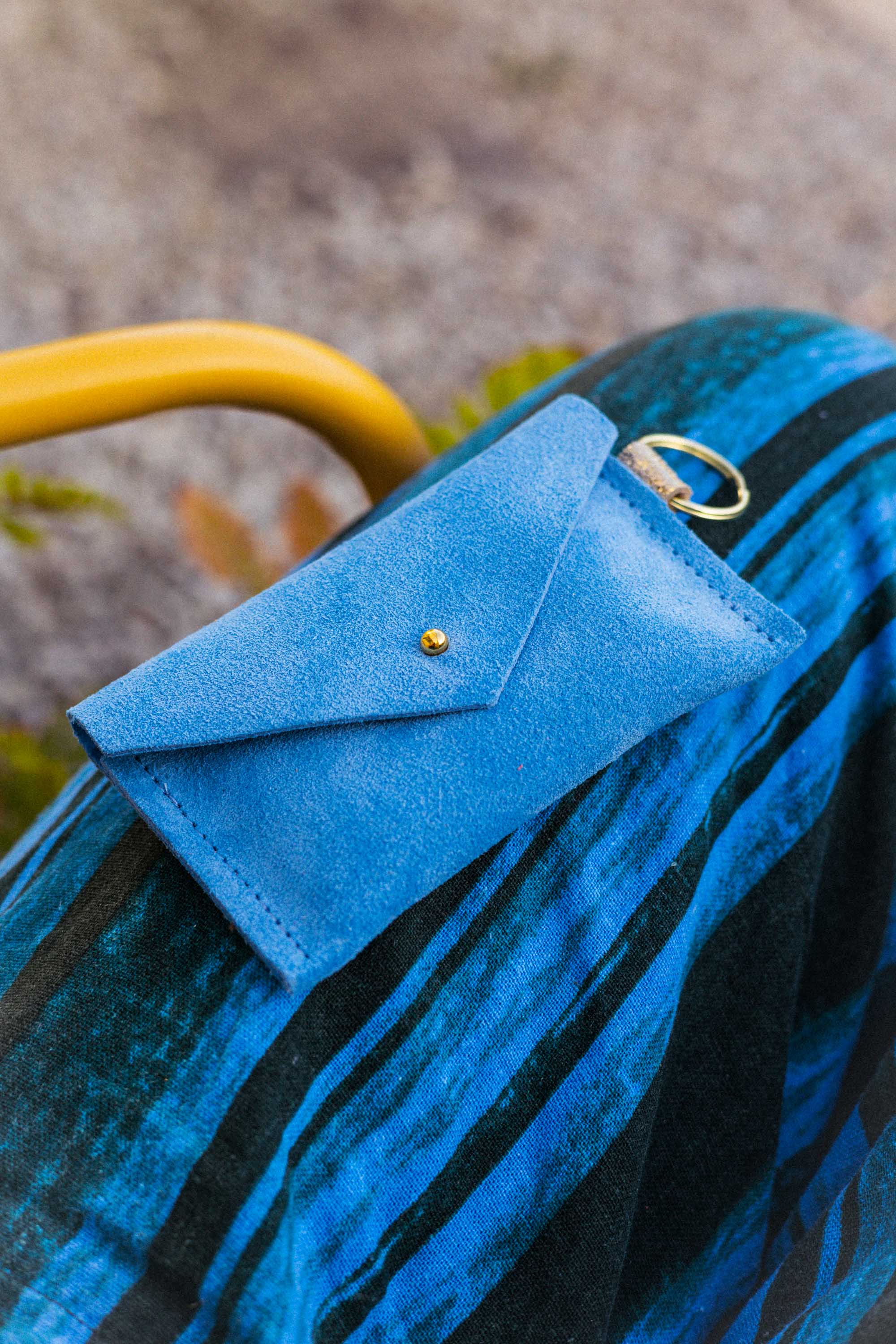 A small sky blue coloured suede envelope-style keyring pouch with a gold button, resting on a blue and black striped patterned fabric.