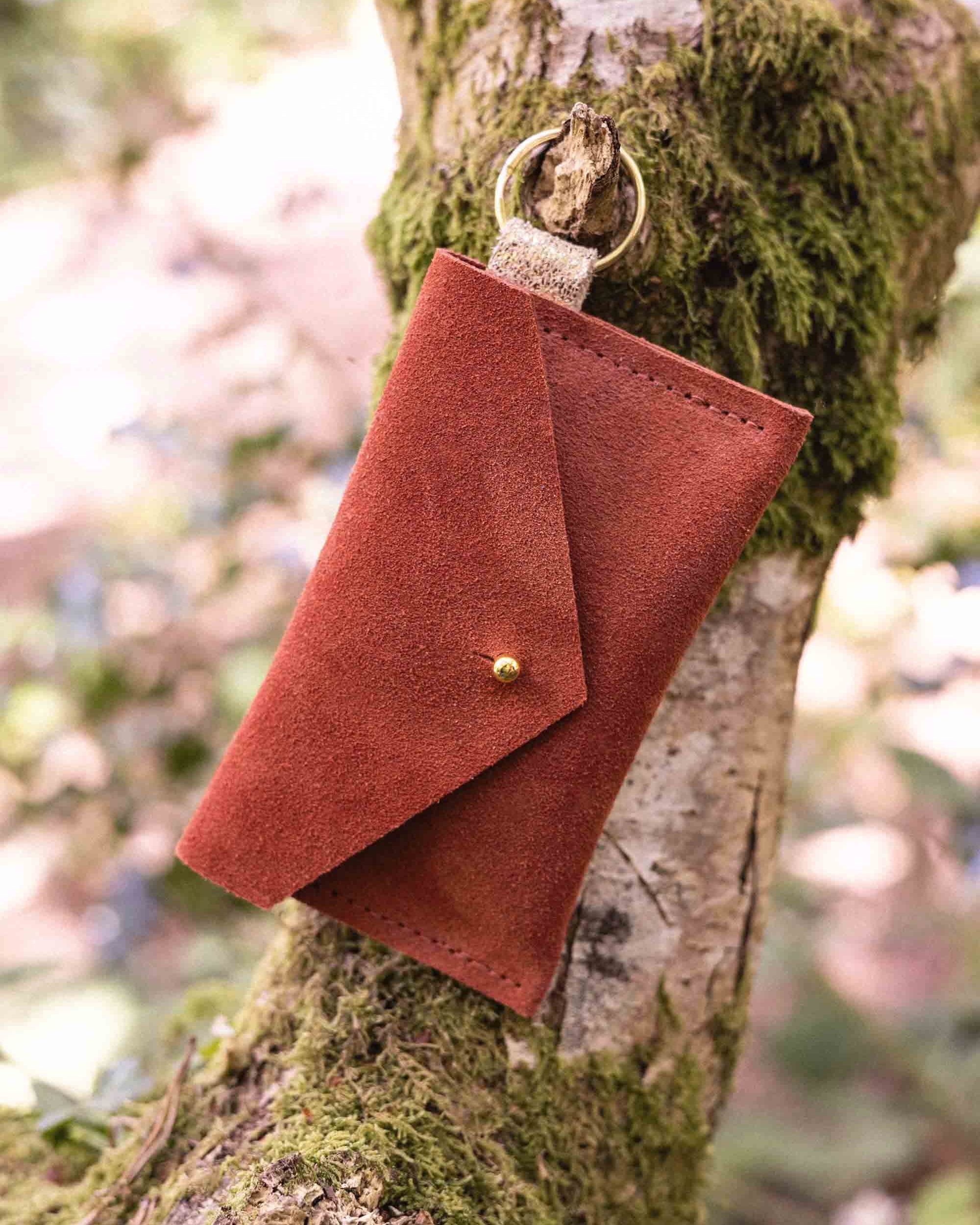A small terracotta coloured suede envelope-style keyring pouch with a gold button, hanging from a branch with a blurred background.