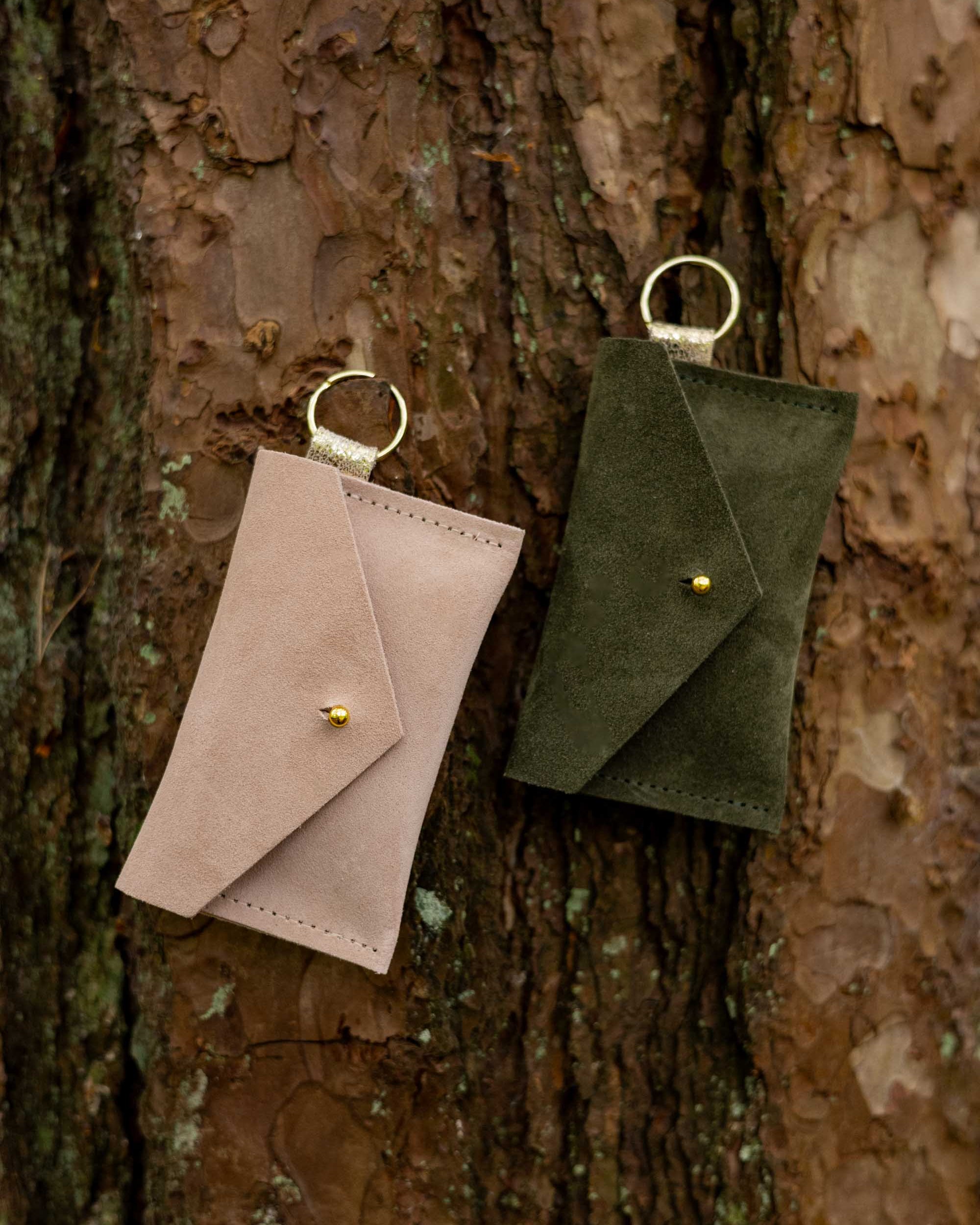 Two small suede envelope-style keyring pouches with a gold button, resting against a textured tree.