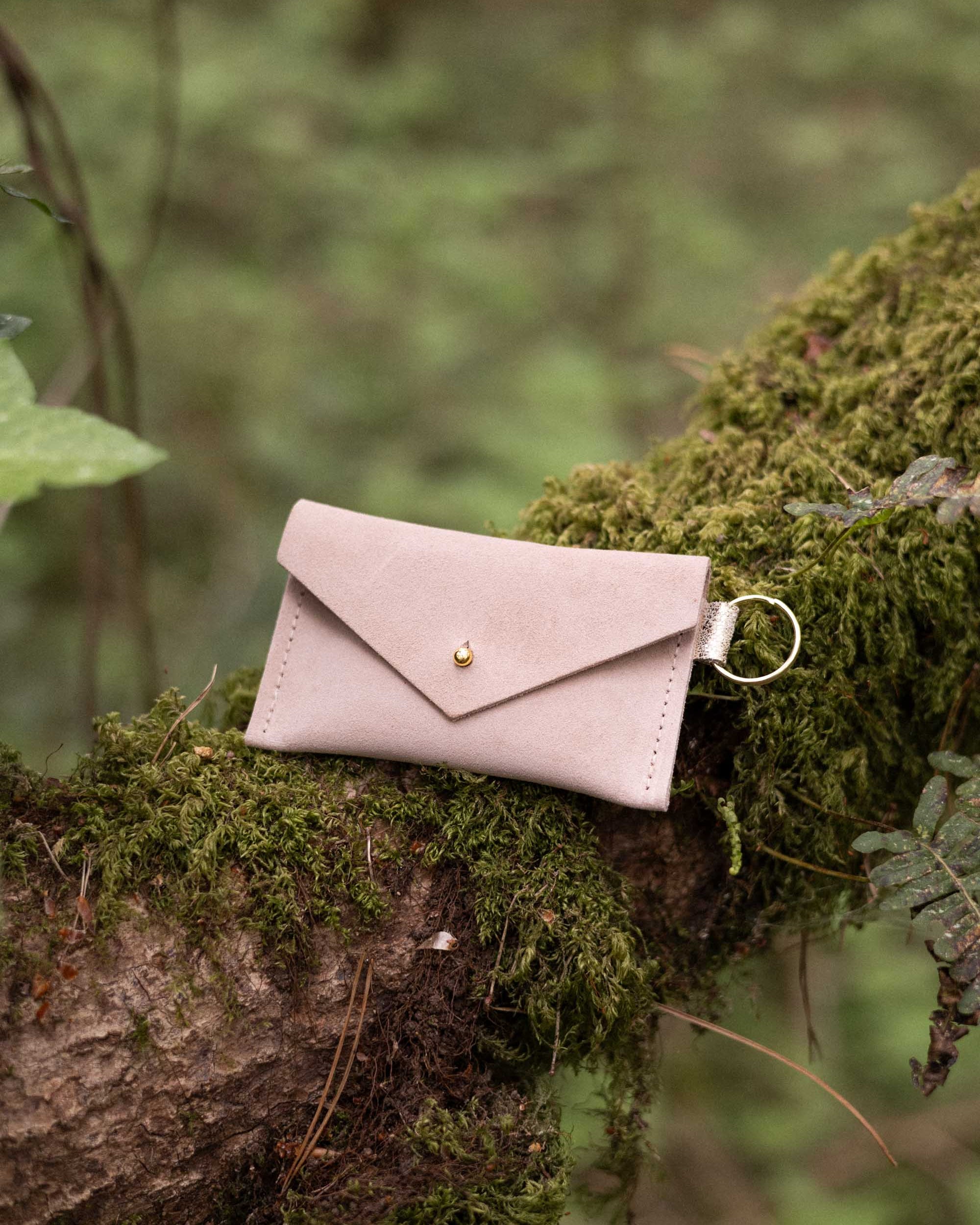 A small warm sand coloured suede envelope-style keyring pouch with a gold button, resting on a branch in a forest.
