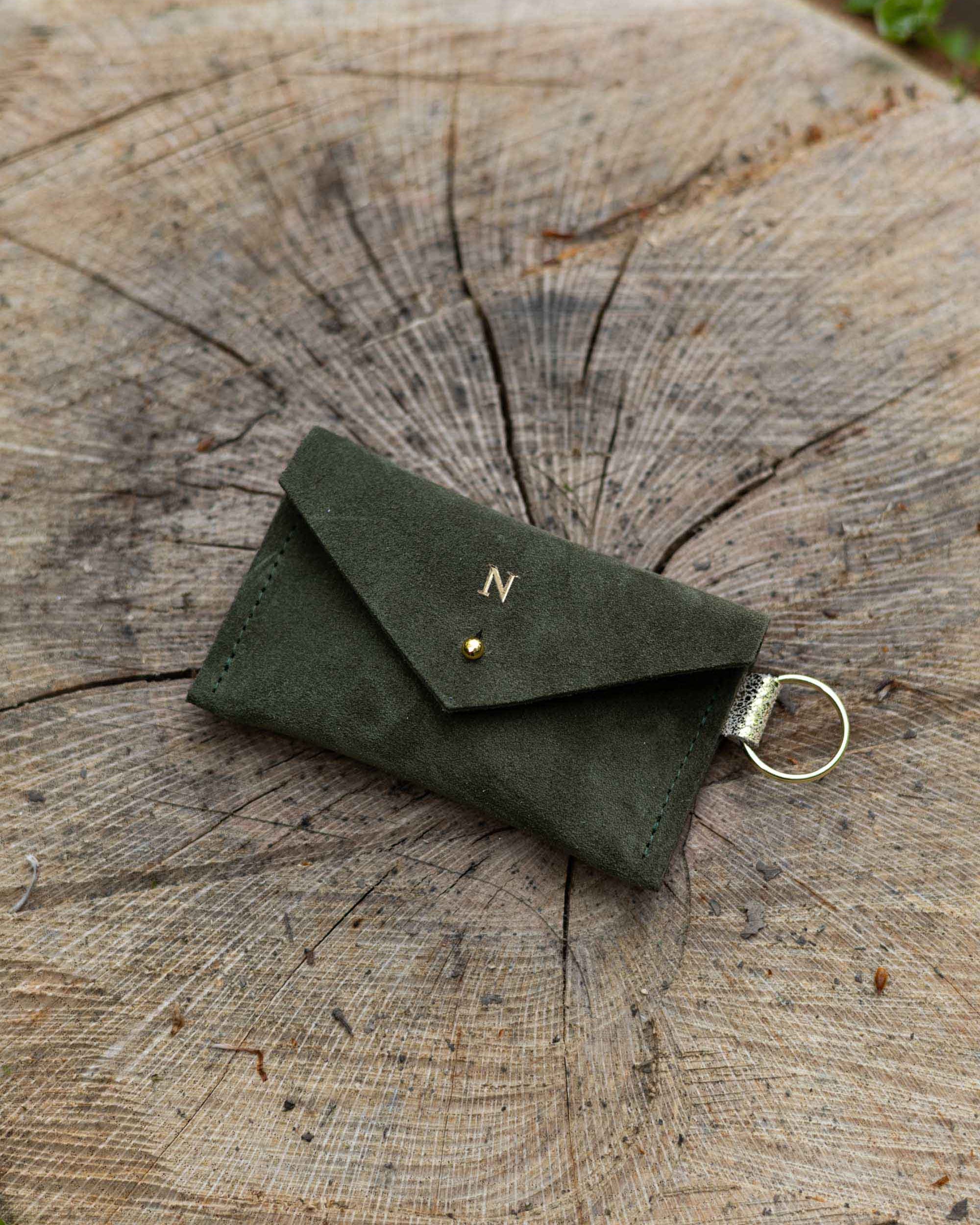 Small soft deep forest coloured suede keyring pouch with a gold foiled initial on the front, resting against a tree.