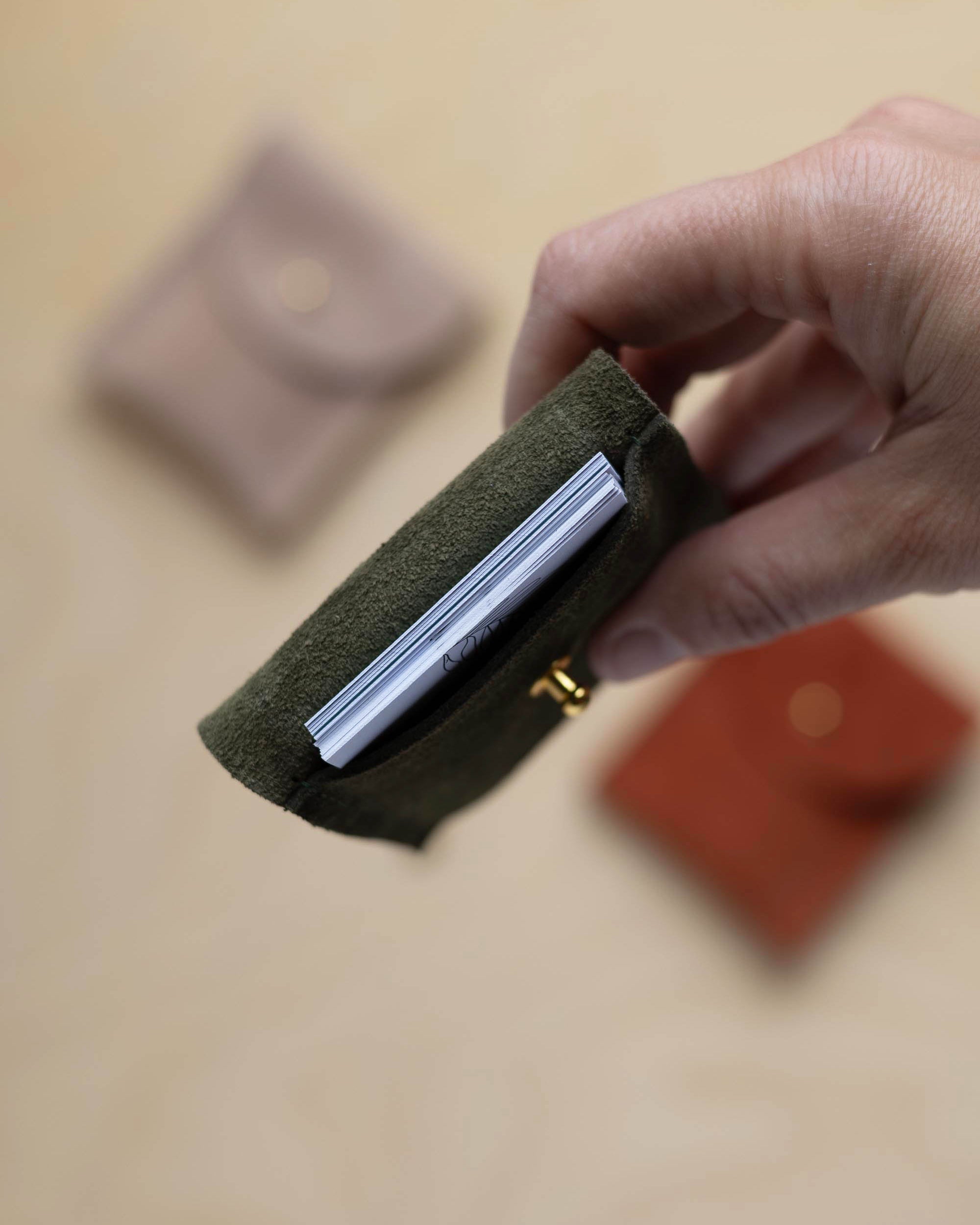 A close up view of a hand holding a deep forest coloured suede pouch with a gold coloured fastening with business cards nestled inside. In the background there are two coloured suede pouches on a light wood grain surface.