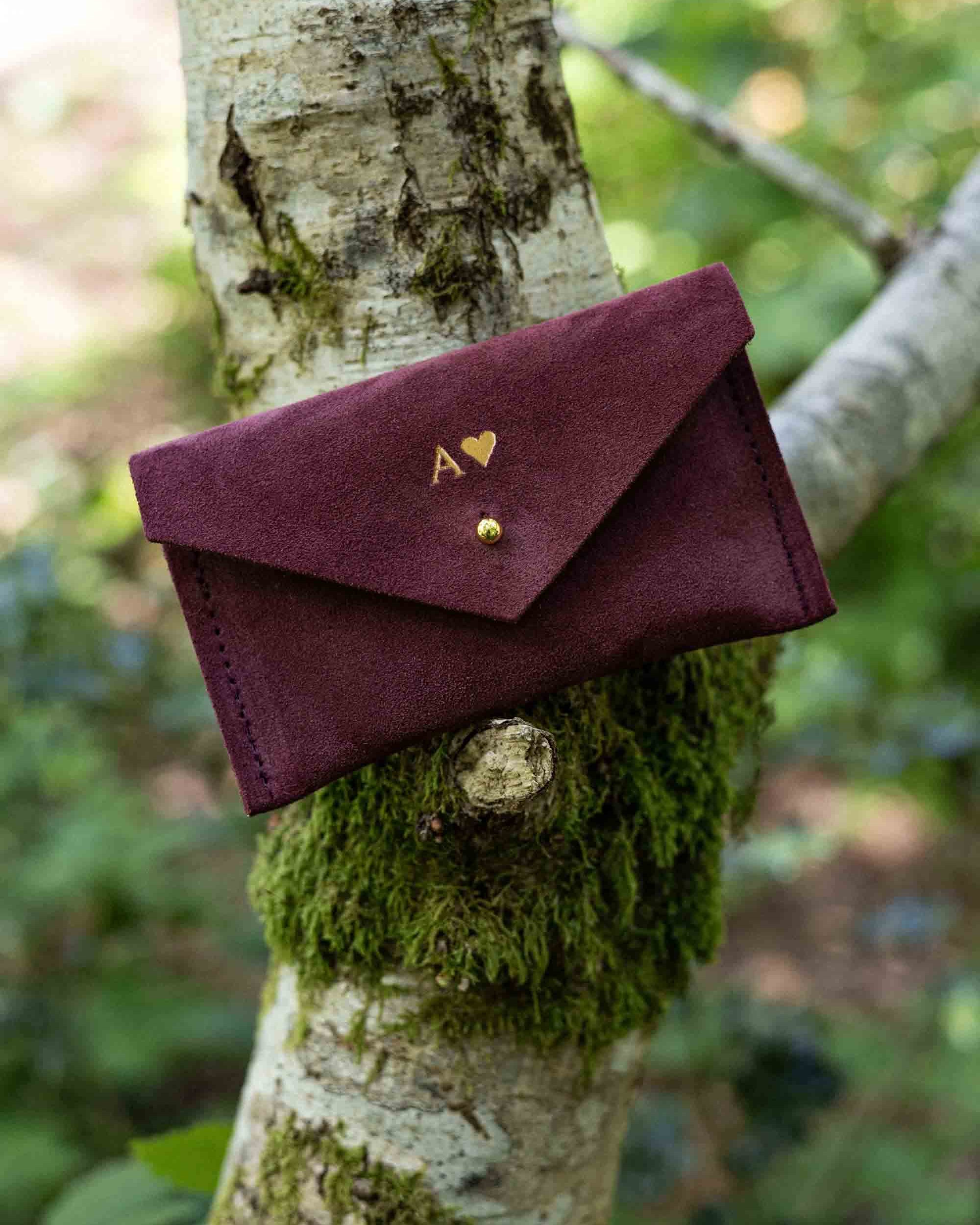 A small burgundy suede pouch with gold foiled initial and heart shape on the front, with a flap and gold button fastening, leaning against a tree branch with an outdoor background.
