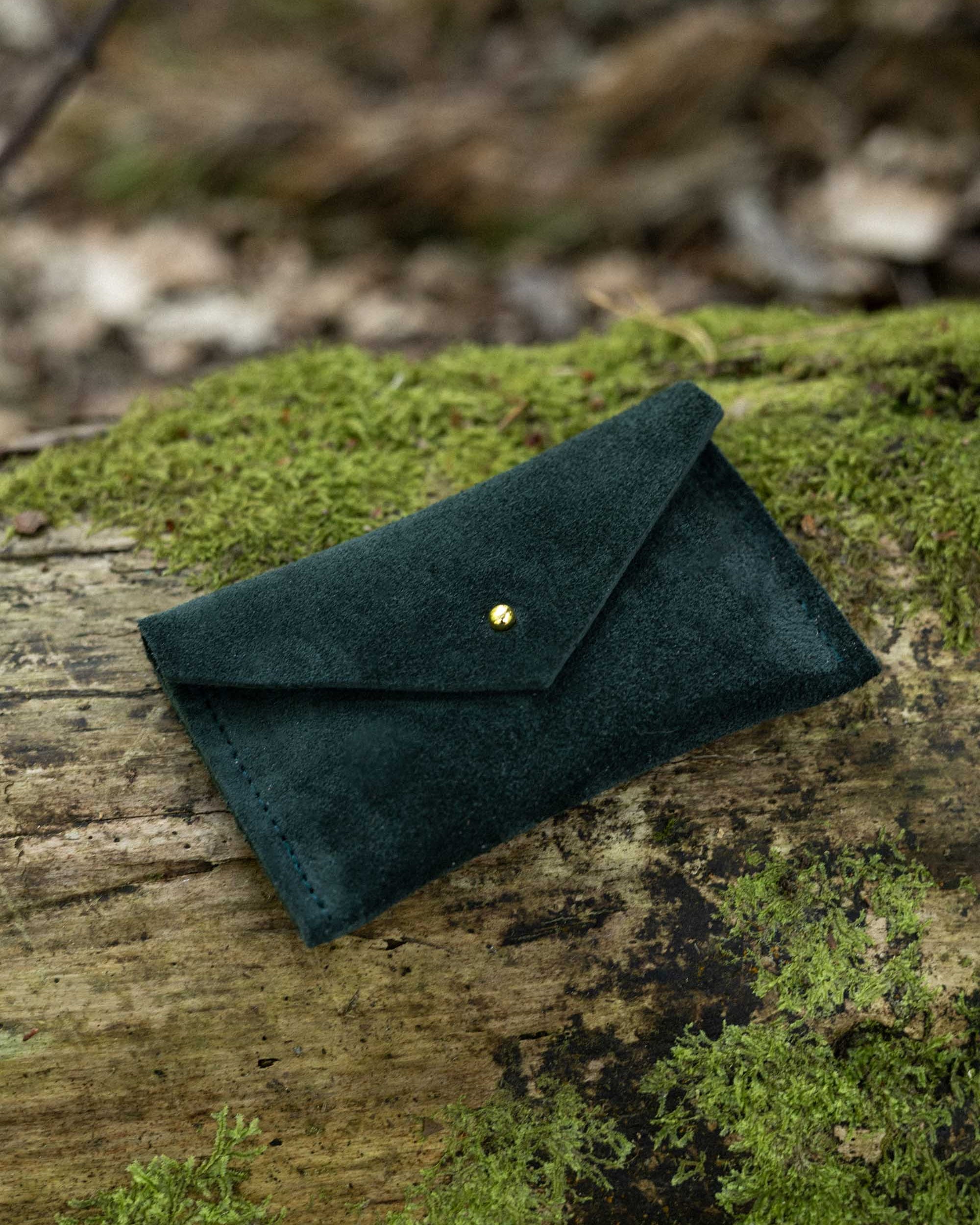 A small deep forest green coloured suede pouch with a flap closure and gold coloured fastening, with an outdoor background.