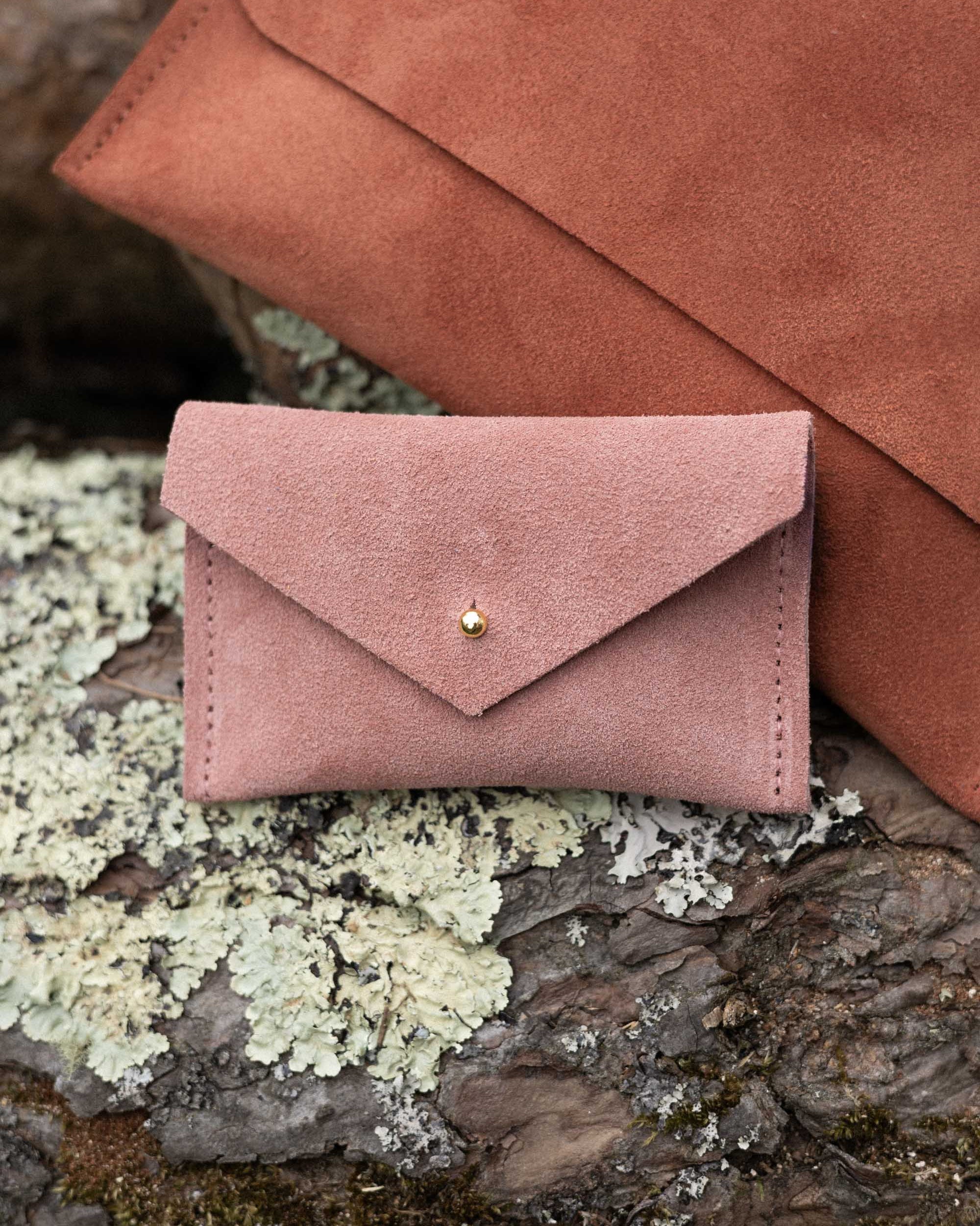 A small dusty rose coloured suede pouch with a flap closure and gold coloured fastening, on a natural wood surface, alongside a terracotta suede clutch bag.