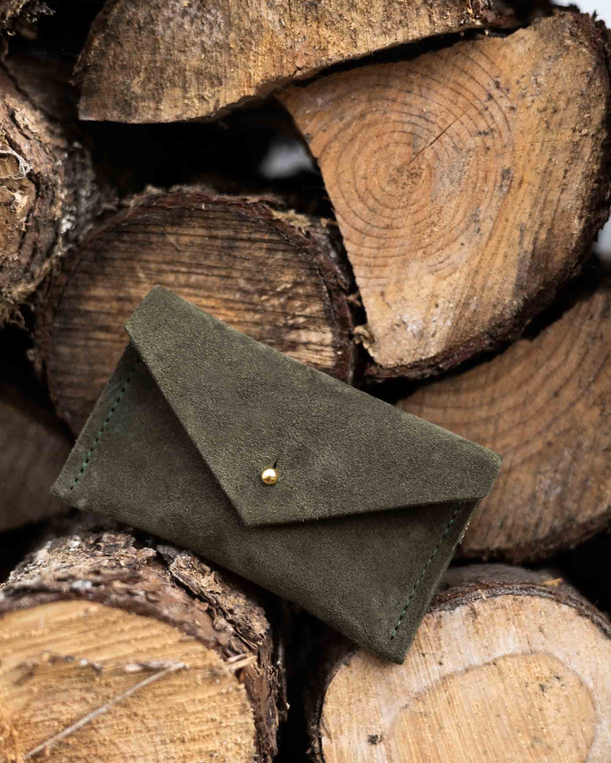 A small soft olive coloured suede pouch with a flap closure and gold coloured fastening, leaning against a wooden natural surface.