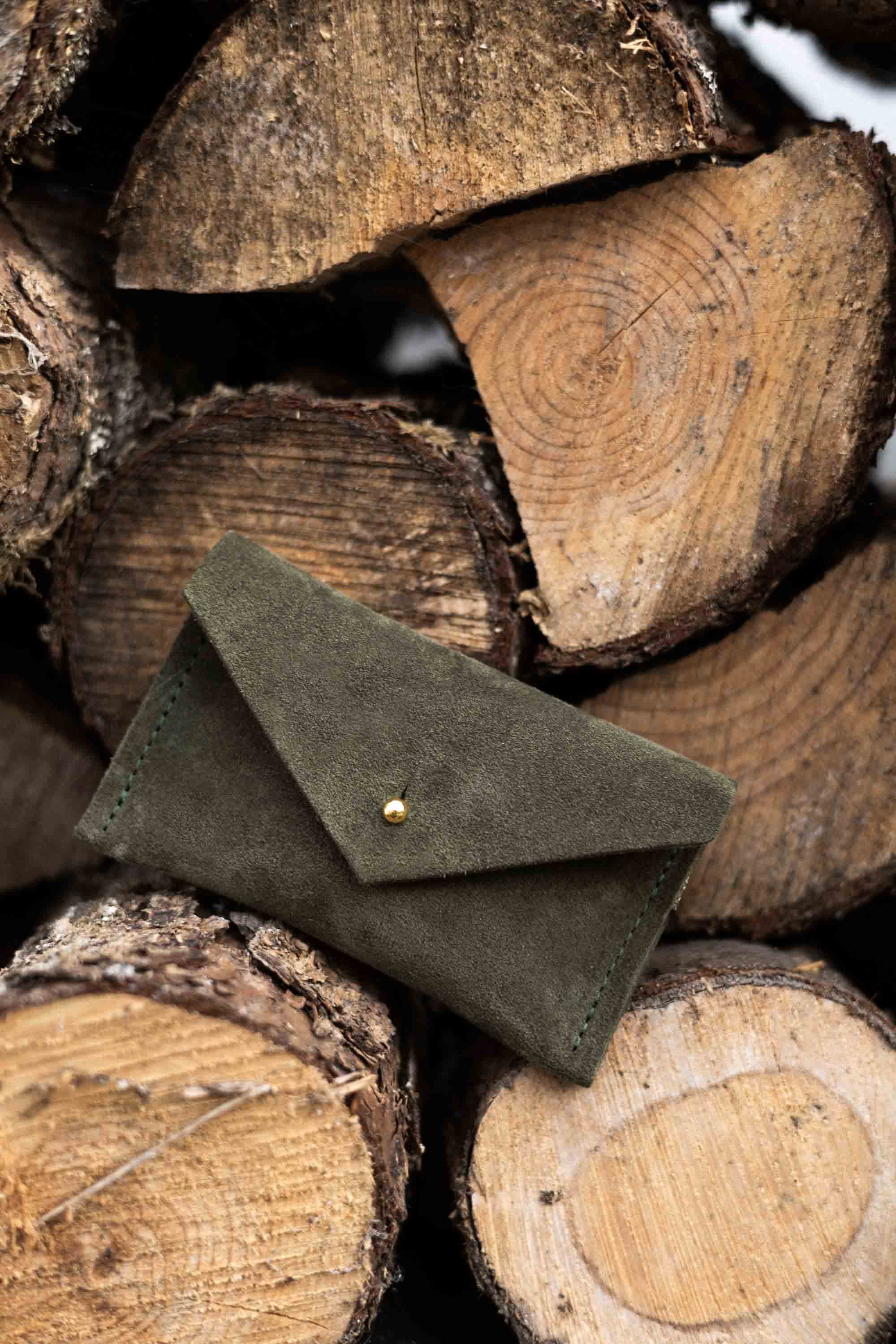 A small soft olive coloured suede pouch with a flap closure and gold coloured fastening, leaning against a wooden natural surface.
