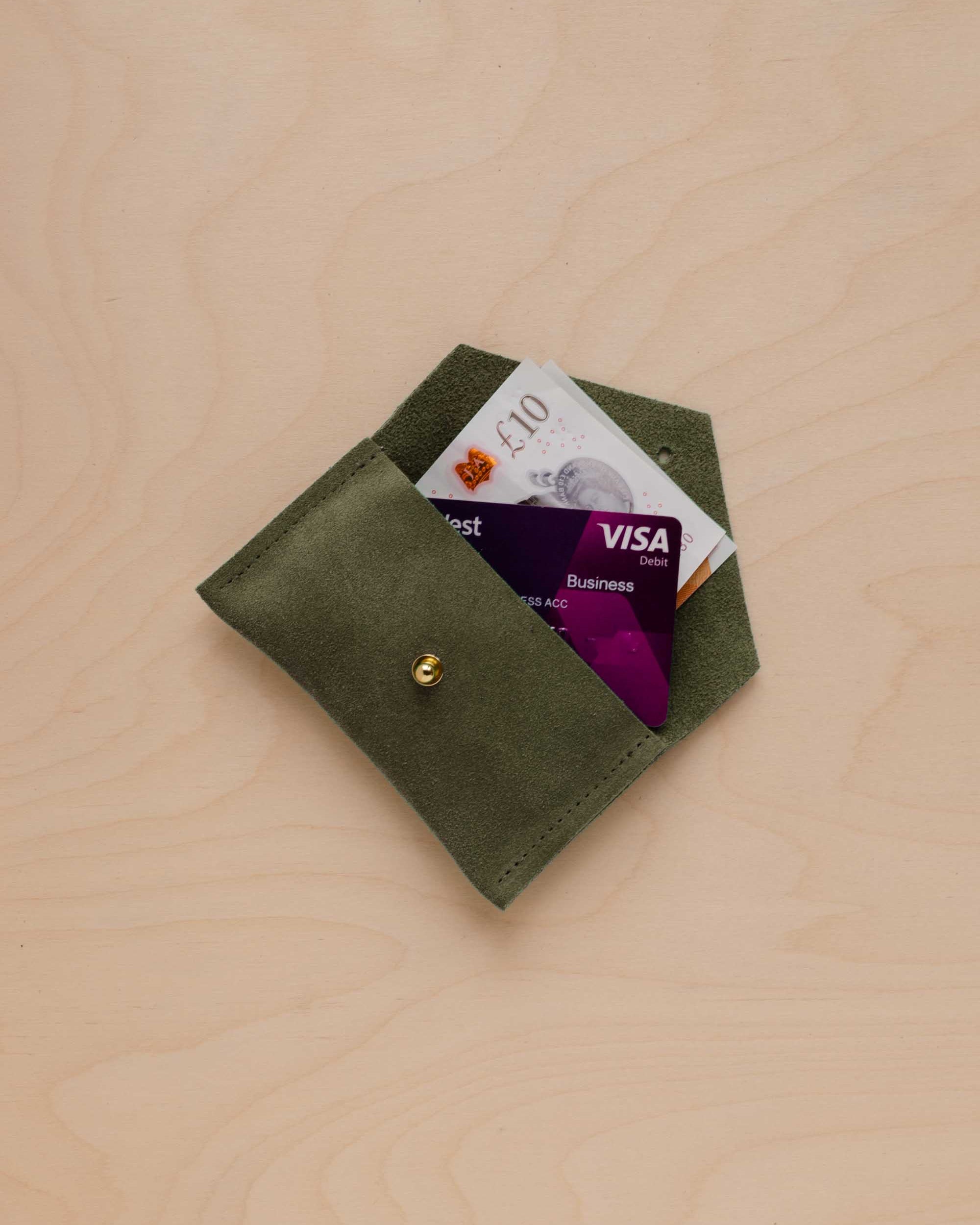 A small soft olive suede pouch with a bank card and bank note nestled inside with a flap and gold button fastening, on a light wood grain surface.
