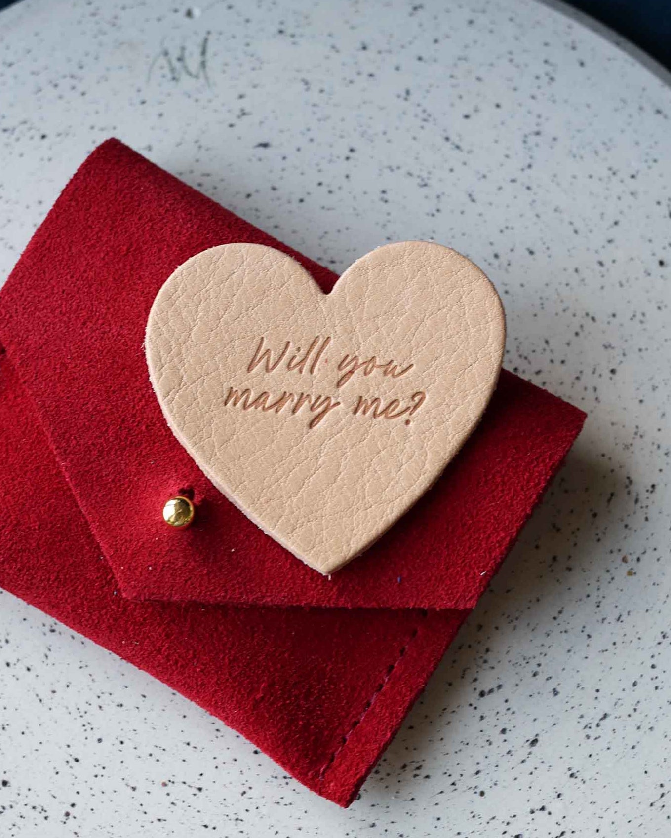 Heart-shaped leather token with 'Will you marry me?' on a red envelope suede pouch.