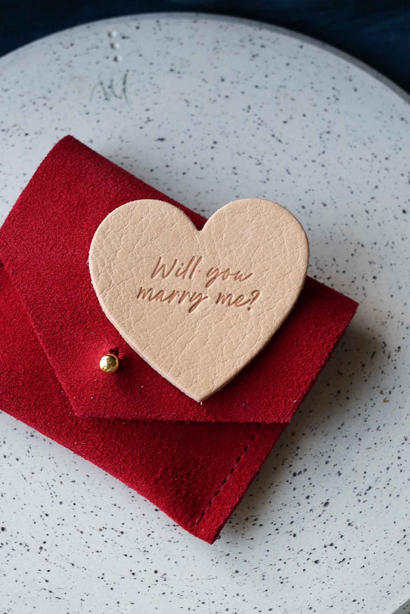 Heart-shaped leather token with 'Will you marry me?' on a red envelope suede pouch.