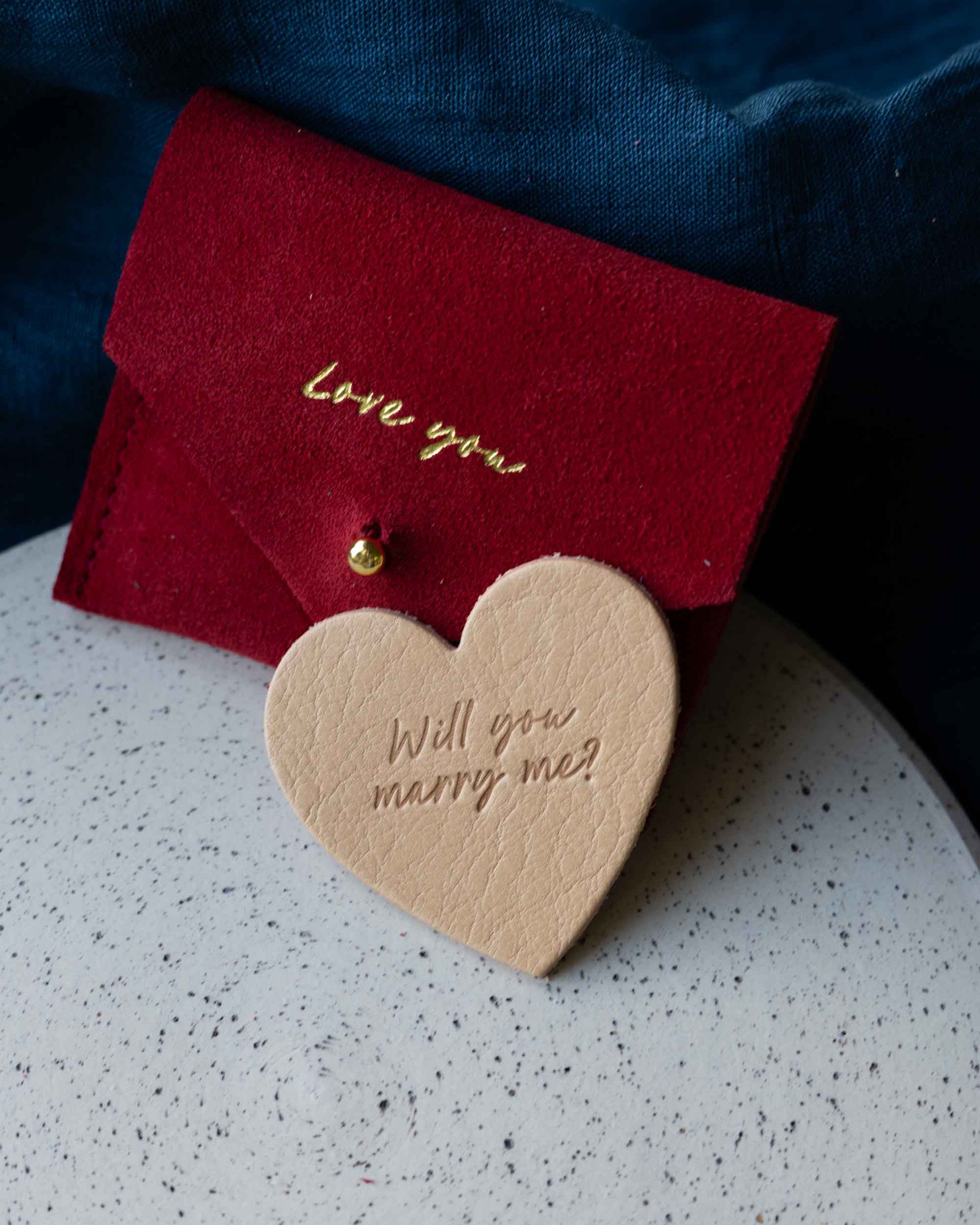 Red card with 'Love you' and heart-shaped wooden tag with 'Will you marry me?' on a speckled surface.