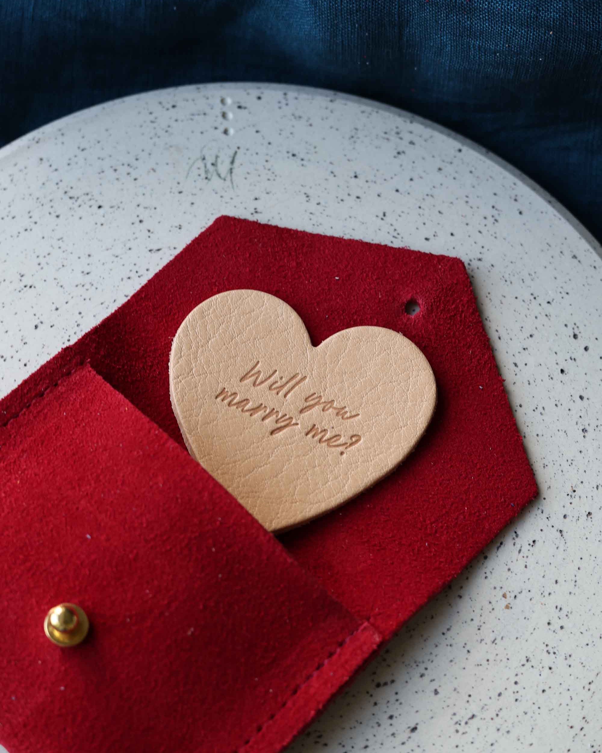 Heart-shaped leather token with 'Will you marry me?' in a red envelope suede pouch.