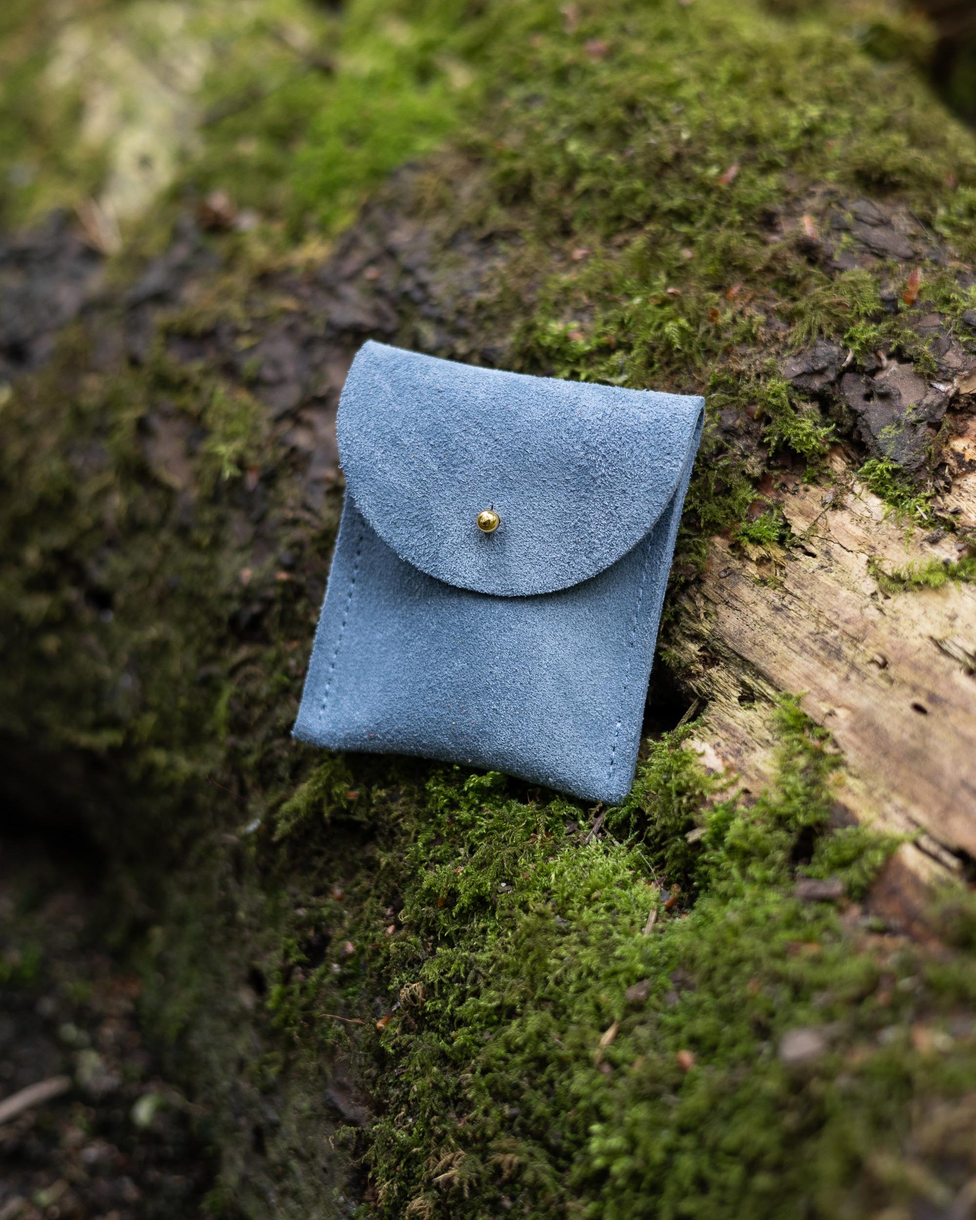 A small dusky blue coloured suede pouch with a gold coloured fastening and flap closure, placed on a natural wooden and moss natural background.