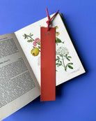 Red leather bookmark with initials on an open book with botanical illustrations against a blue background