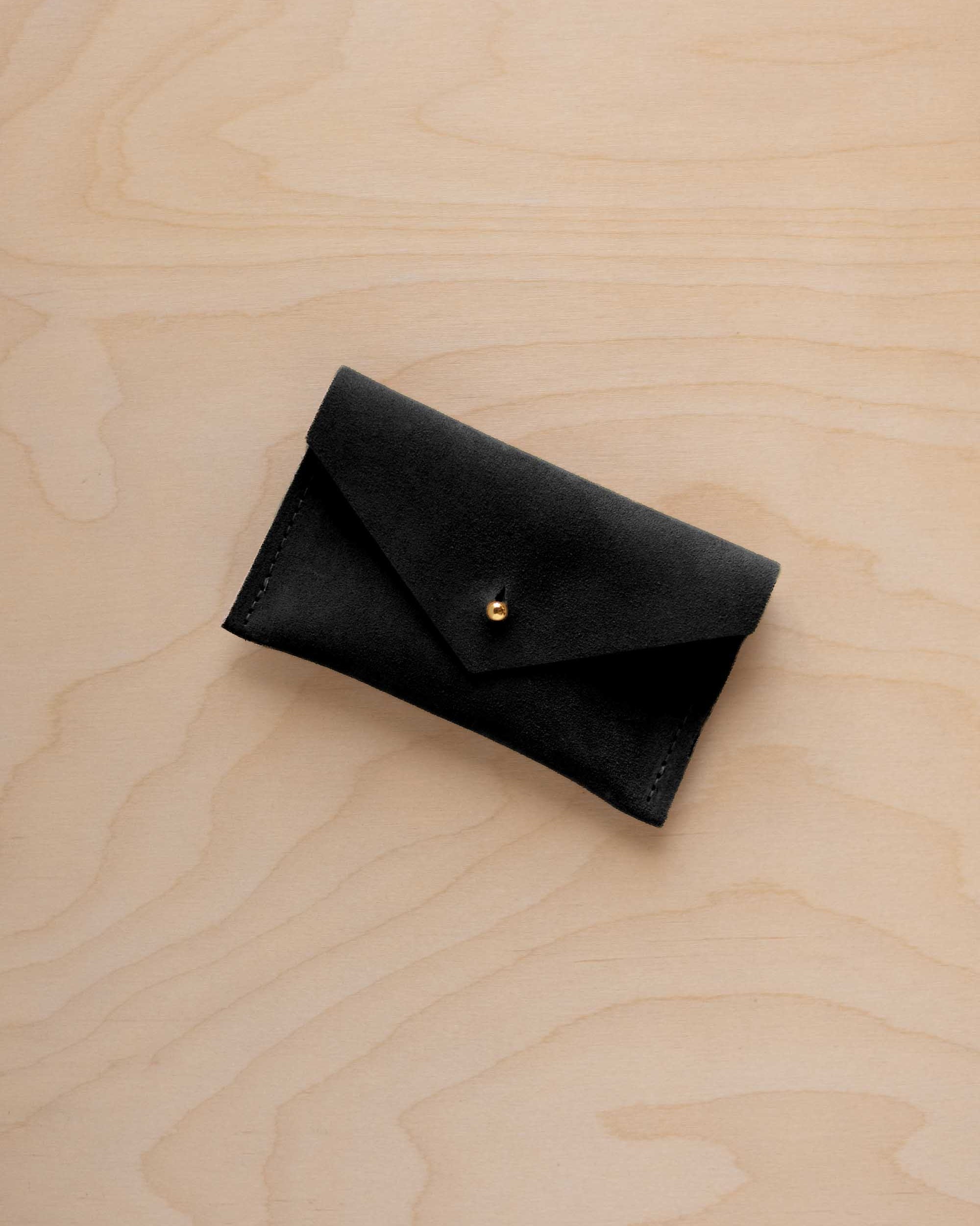 A small black coloured suede pouch with a flap closure and gold coloured fastening, on a light wood grain surface.