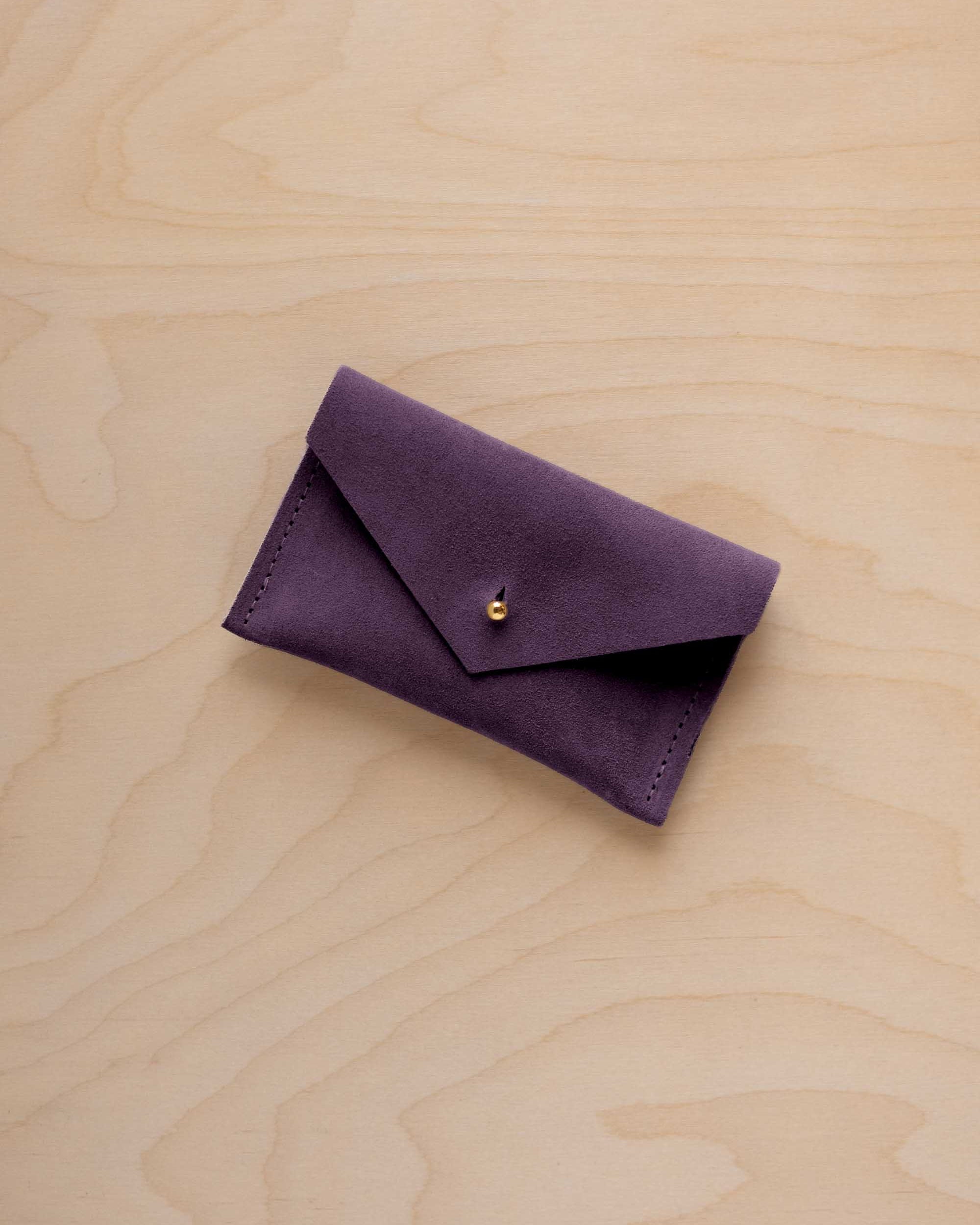 A small blackcurrant coloured suede pouch with a flap closure and gold coloured fastening, on a light wood grain surface.