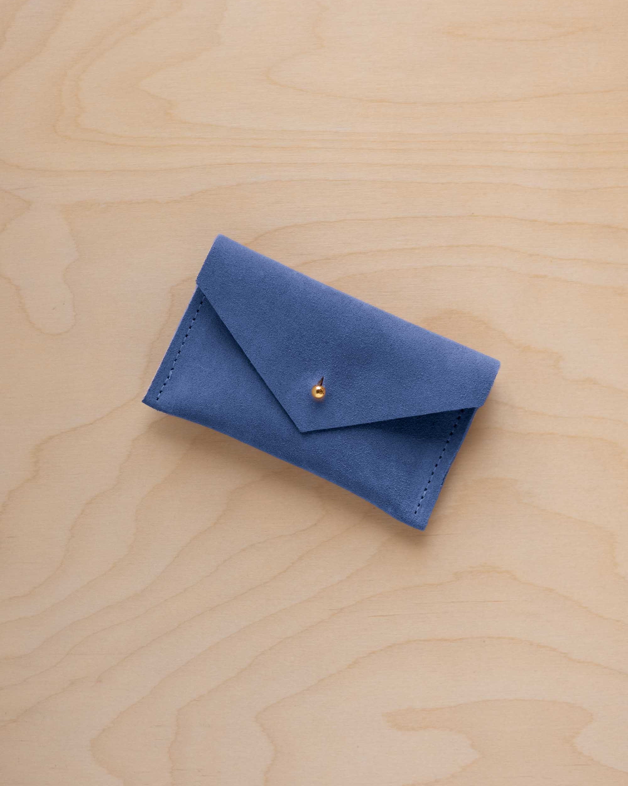 A small cobalt blue coloured suede pouch with a flap closure and gold coloured fastening, on a light wood grain surface.