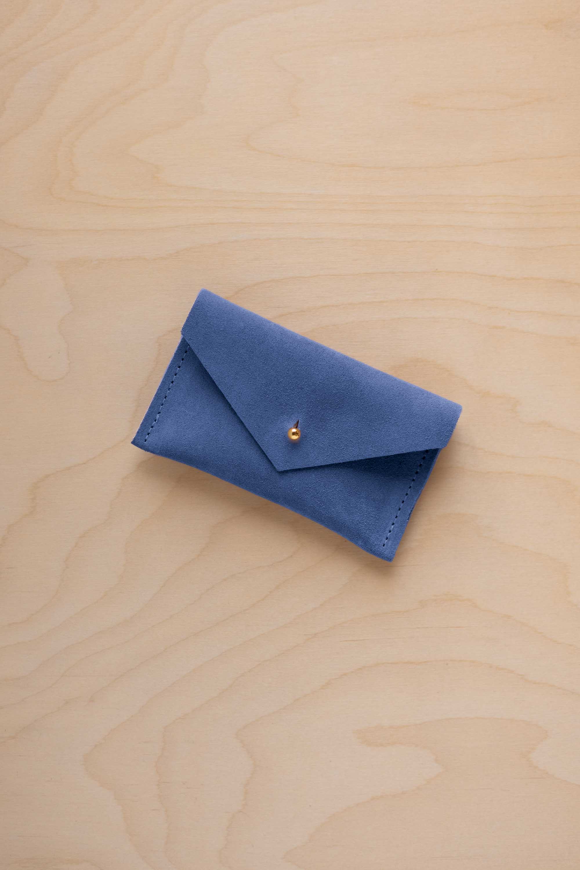 A small cobalt blue coloured suede pouch with a flap closure and gold coloured fastening, on a light wood grain surface.