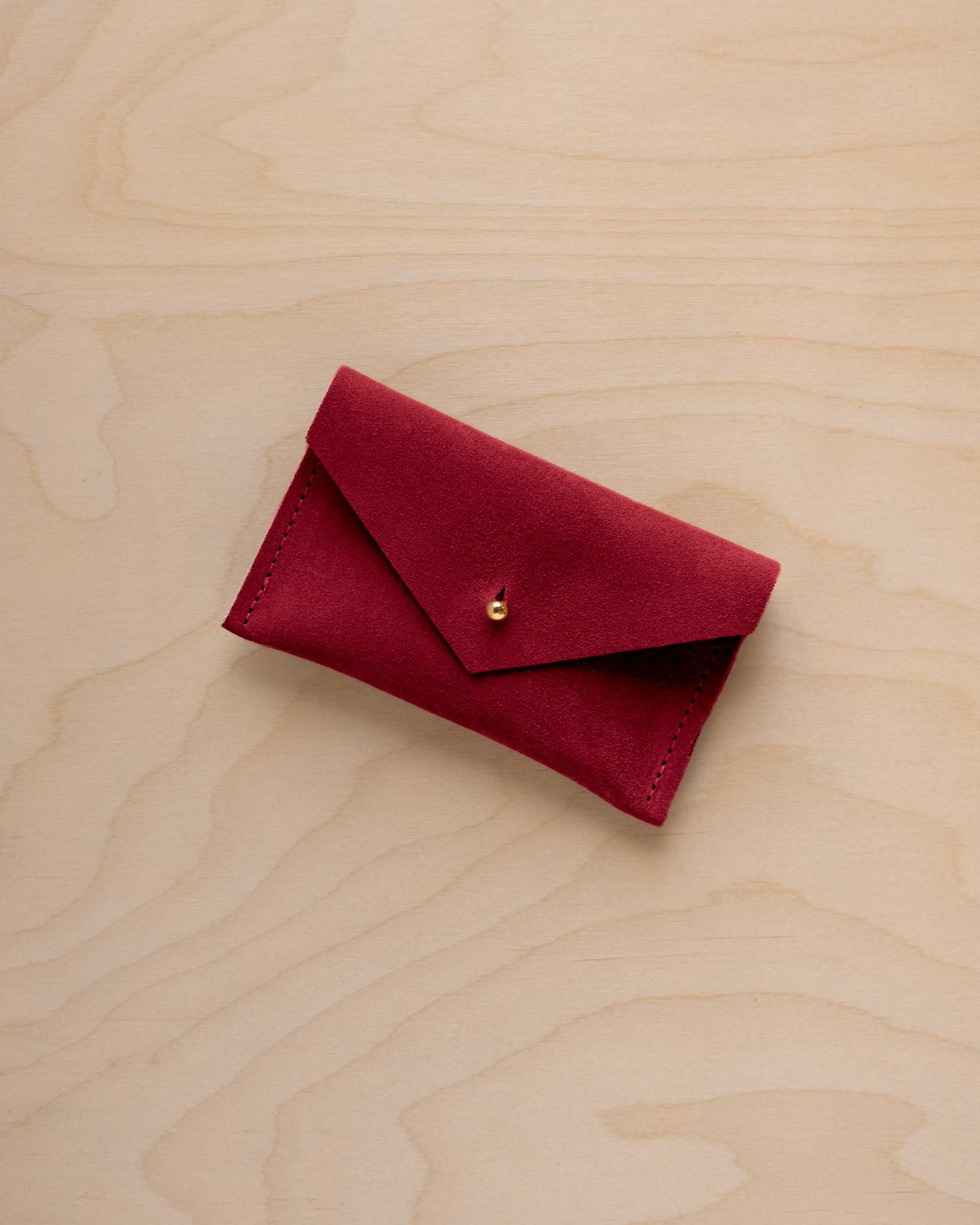 A small crimson red coloured suede pouch with a flap closure and gold coloured fastening, on a light wood grain surface.