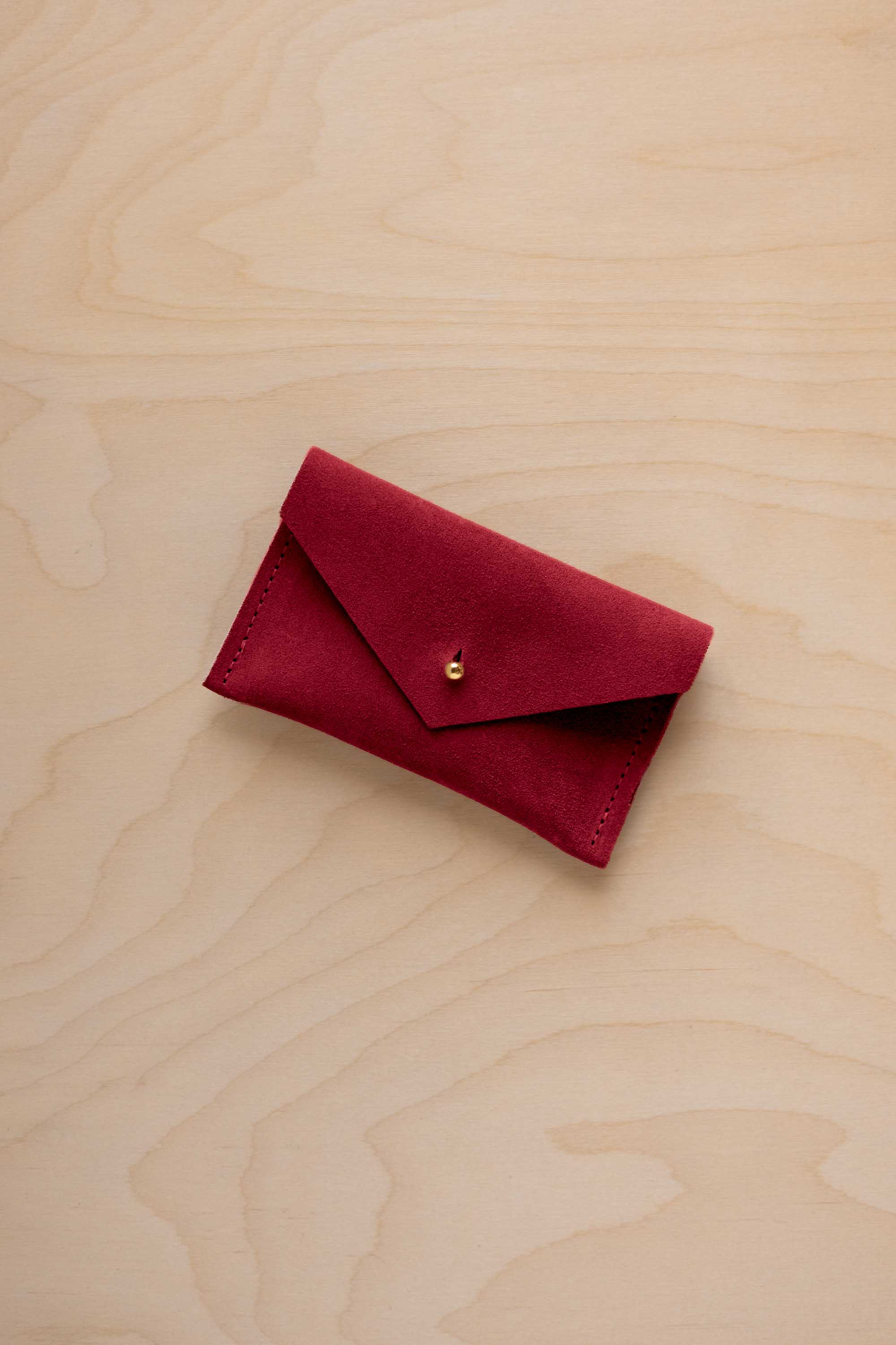 A small crimson red coloured suede pouch with a flap closure and gold coloured fastening, on a light wood grain surface.