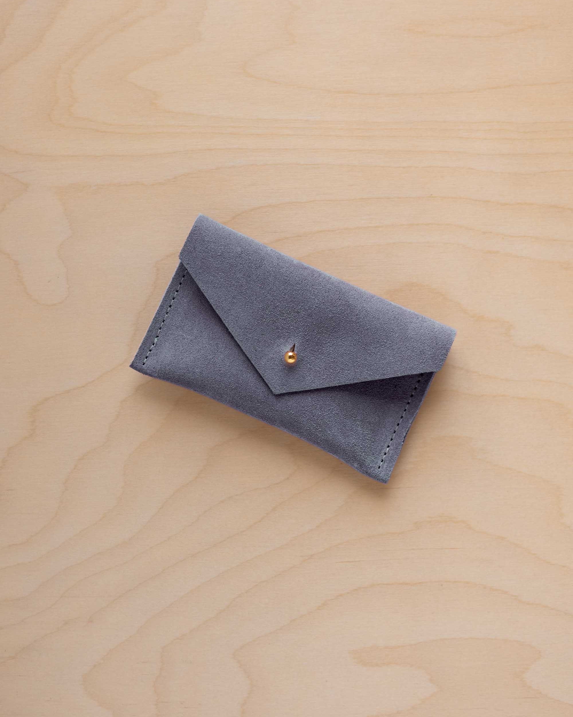 A small dusty blue coloured suede pouch with a flap closure and gold coloured fastening, on a light wood grain surface.