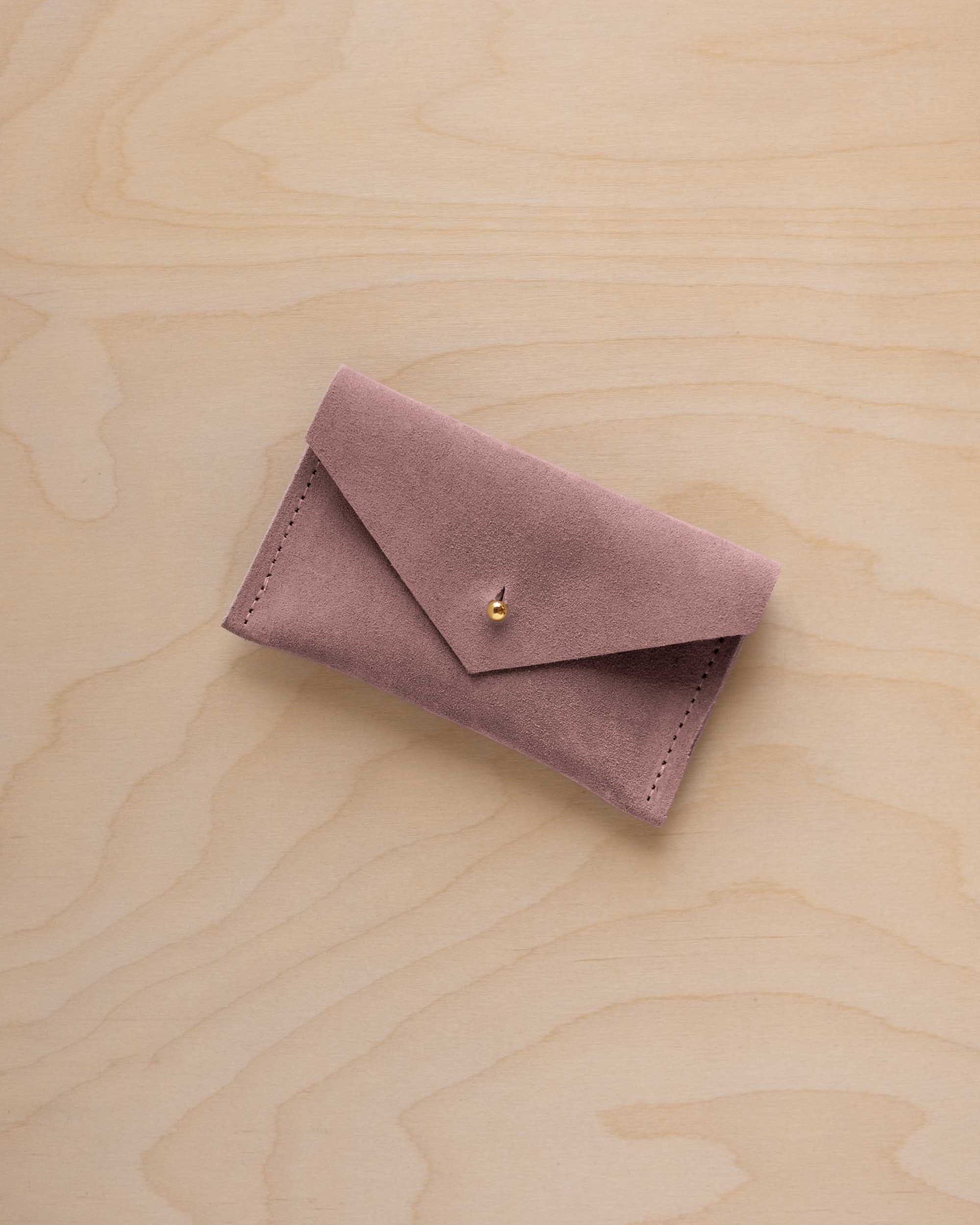 A small dusty mauve coloured suede pouch with a flap closure and gold coloured fastening, on a light wood grain surface.